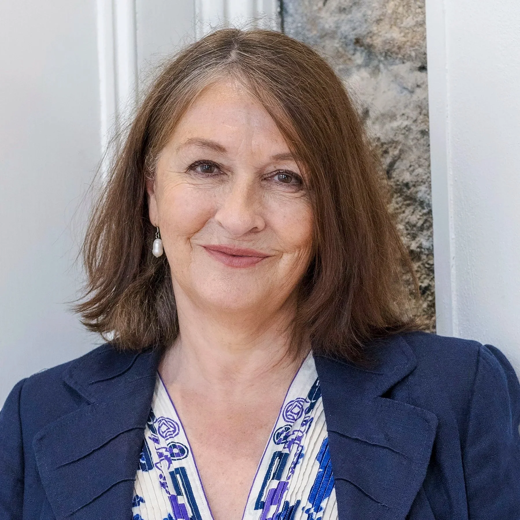 Close-up portrait of a woman with wavy, shoulder-length brown hair, wearing pearl earrings, a patterned shirt, and a dark blazer, smiling at the camera.