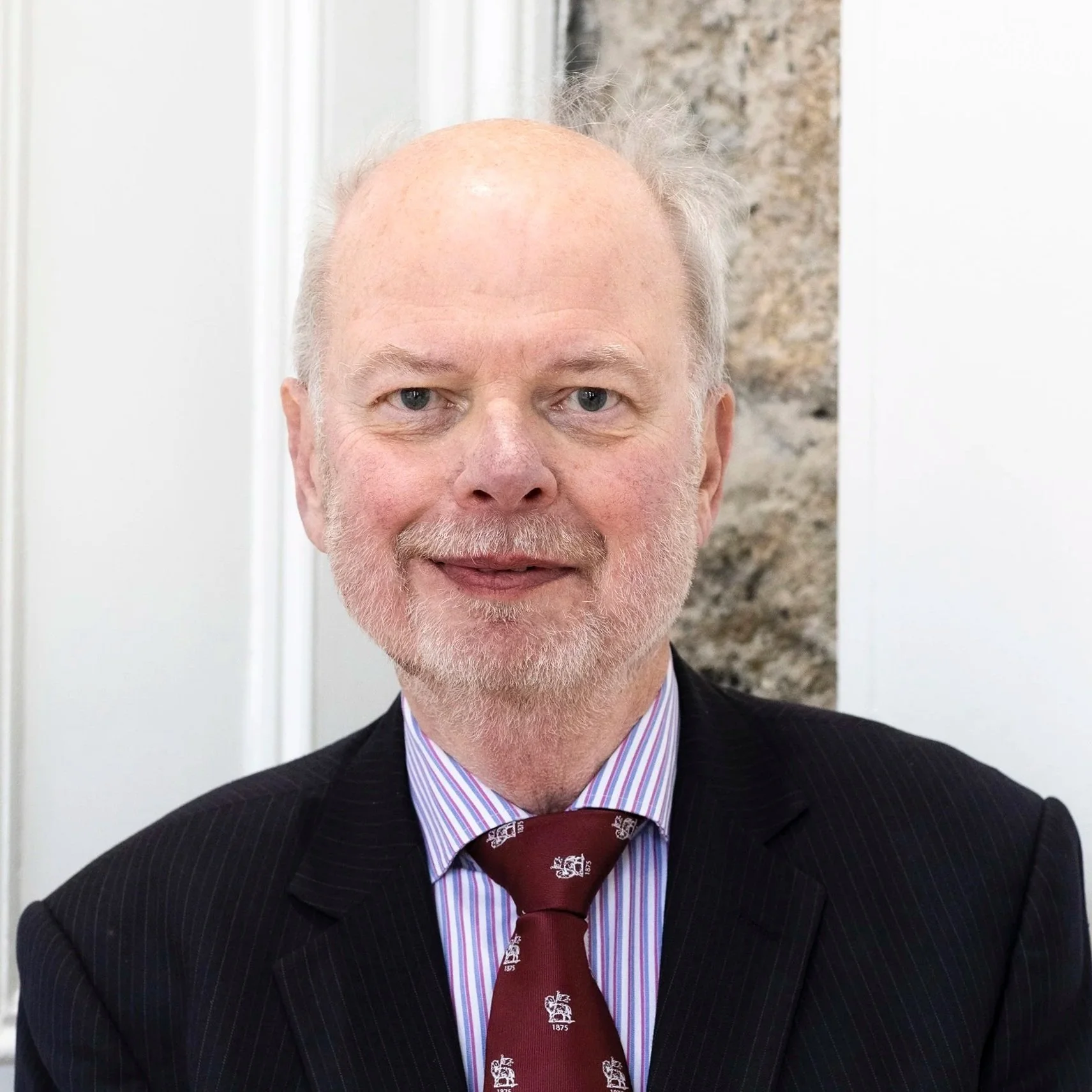 Close-up of a man with light skin, blue eyes, and a beard, wearing a black suit, striped dress shirt, and a maroon tie with white emblem, standing indoors with a white and textured wall background.