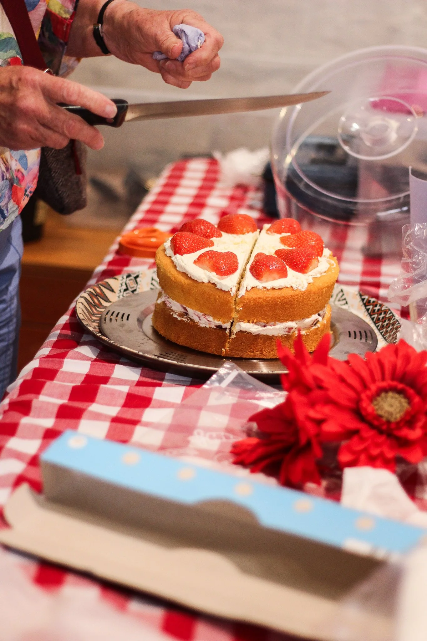 A person cutting a strawberry-topped cake with a large knife on a red and white checkered tablecloth, with a flower in the foreground and a plastic container in the background.