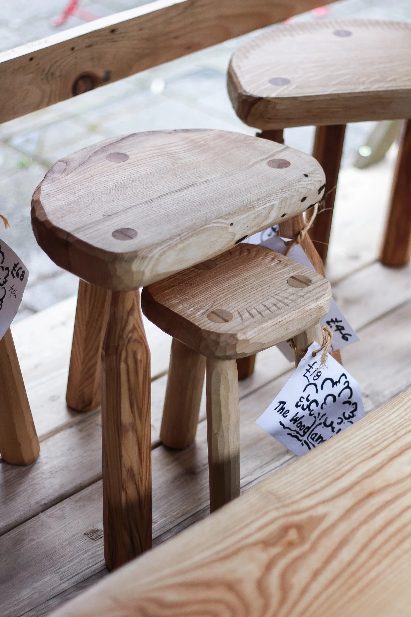 Close-up of wooden stools made of light-colored wood with visible grain, secured with round wooden pegs, arranged in a store display on a wooden table.