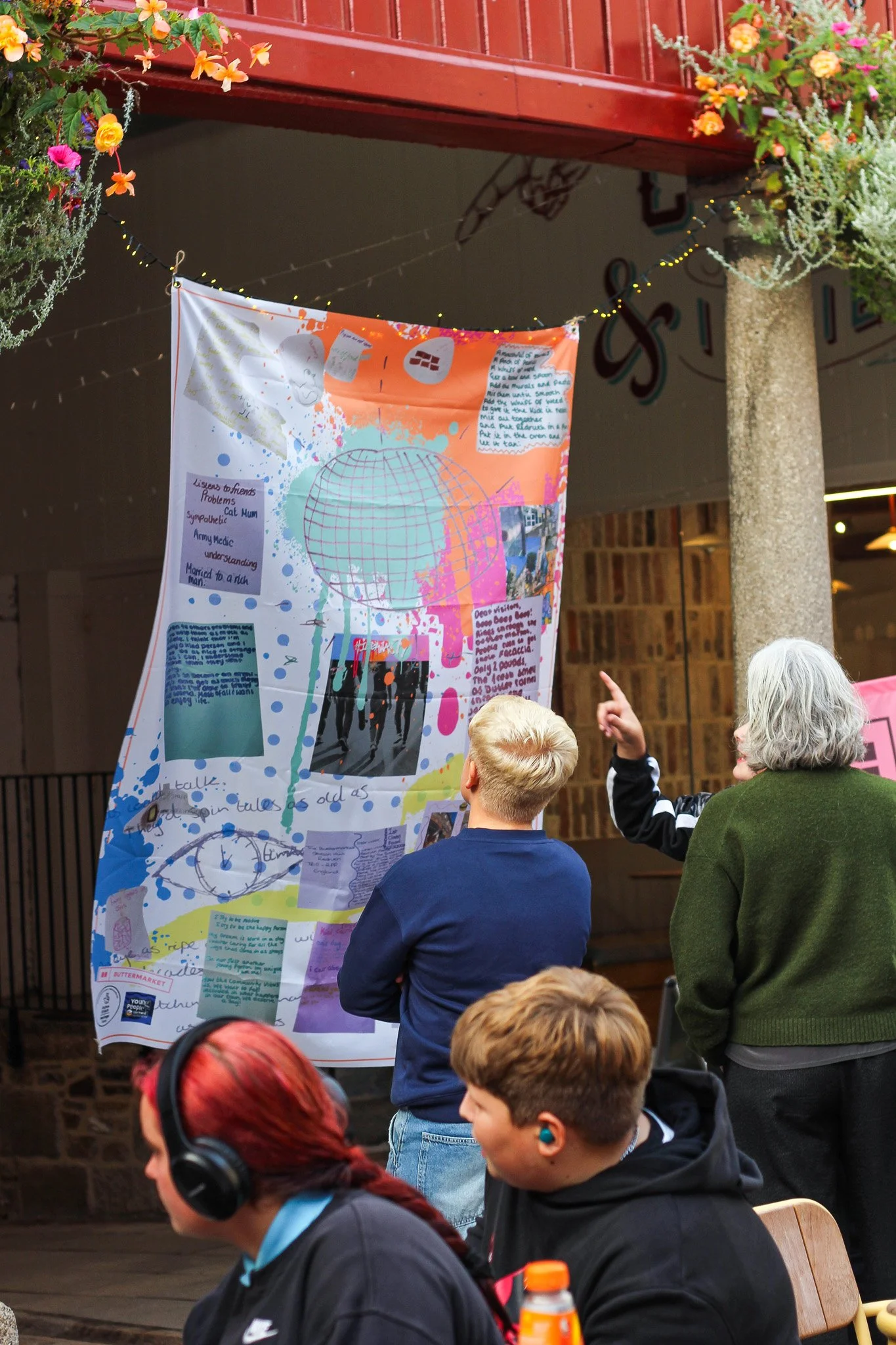People gathered around a colorful poster hanging from a string, with a red railing and hanging flower baskets above. Children and adults are looking at the poster, which contains handwritten notes, drawings, and images.
