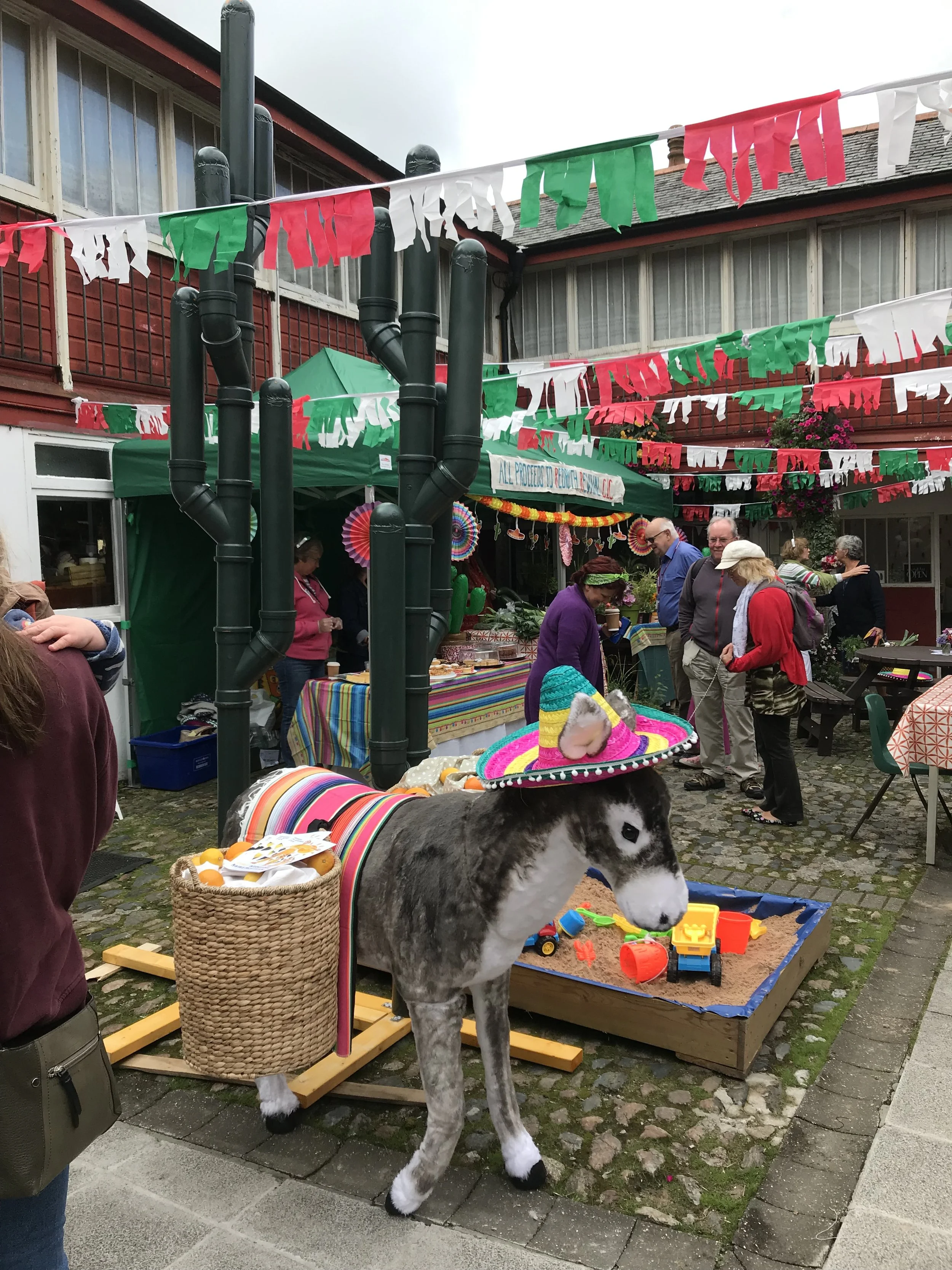 A lively outdoor market or festival scene with colorful decorations and several people browsing and socializing. A small horse or pony with a colorful sombrero is visible near a sandpit filled with toy trucks and toys.