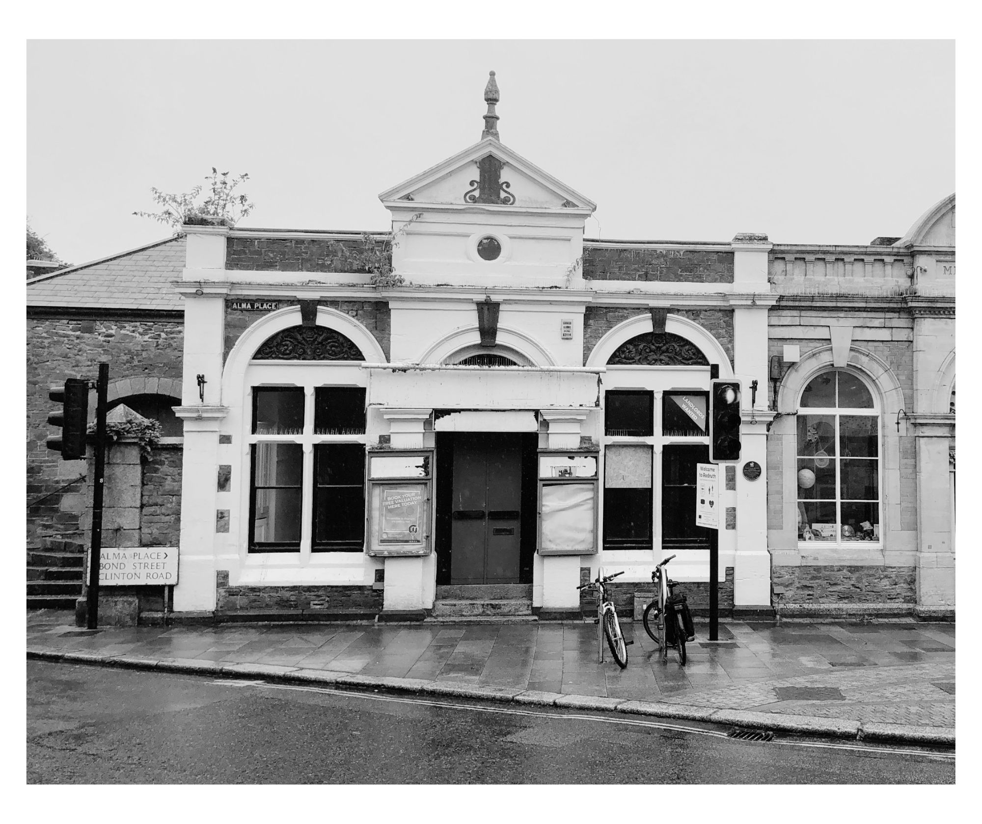 Black and white photo of an old building with arched windows and a decorative triangular pediment on top, with a street and bicycles in front.
