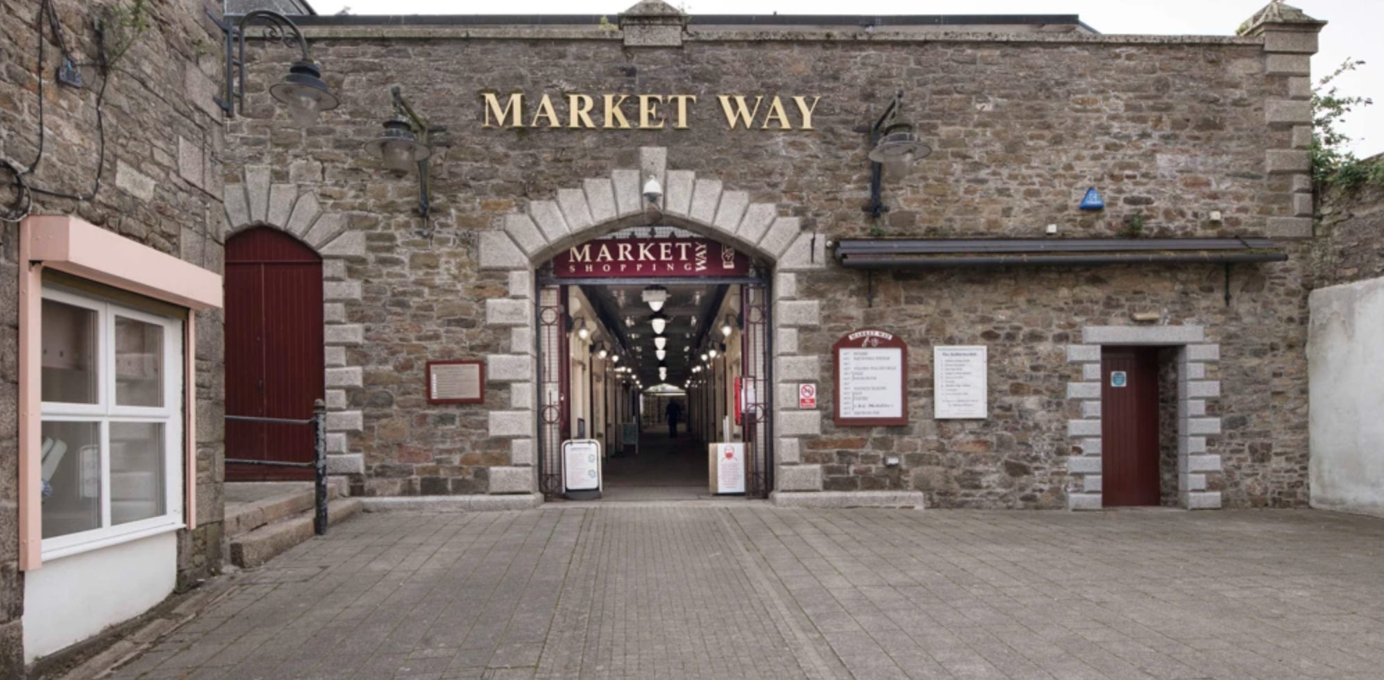 Entrance to a market shopping area with a stone brick facade, dark metal gates, and illuminated signs reading 'MARKET WAY' and 'MARKET SHOPPING'.
