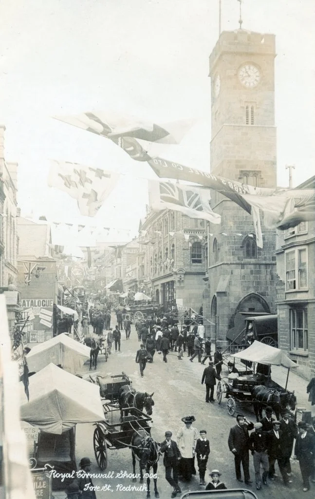 A busy 1906 street scene at the Royal Cornwall Show in Redruth, featuring horse-drawn carriages, pedestrians, storefronts, period clothing, American and British flags, and a prominent clock tower.
