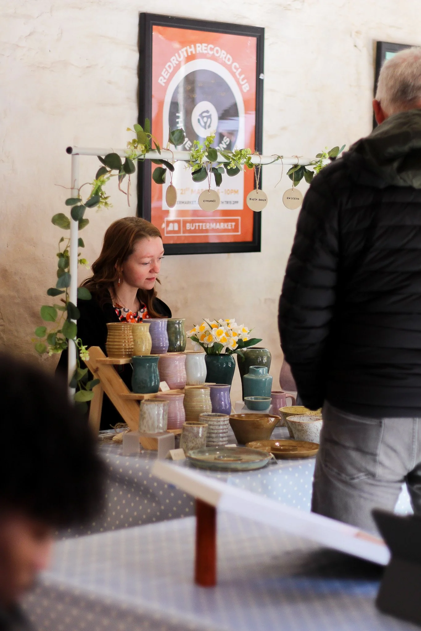 A display table of colorful ceramic vases and bowls at an indoor market or craft fair, with a woman sitting behind the table and a man standing in front of it, and a framed poster on the wall behind them.