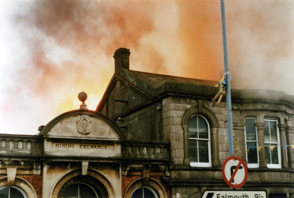 A stone building with a sign reading 'Mining Exchange' is on fire, with large orange flames and thick smoke rising from the roof.
