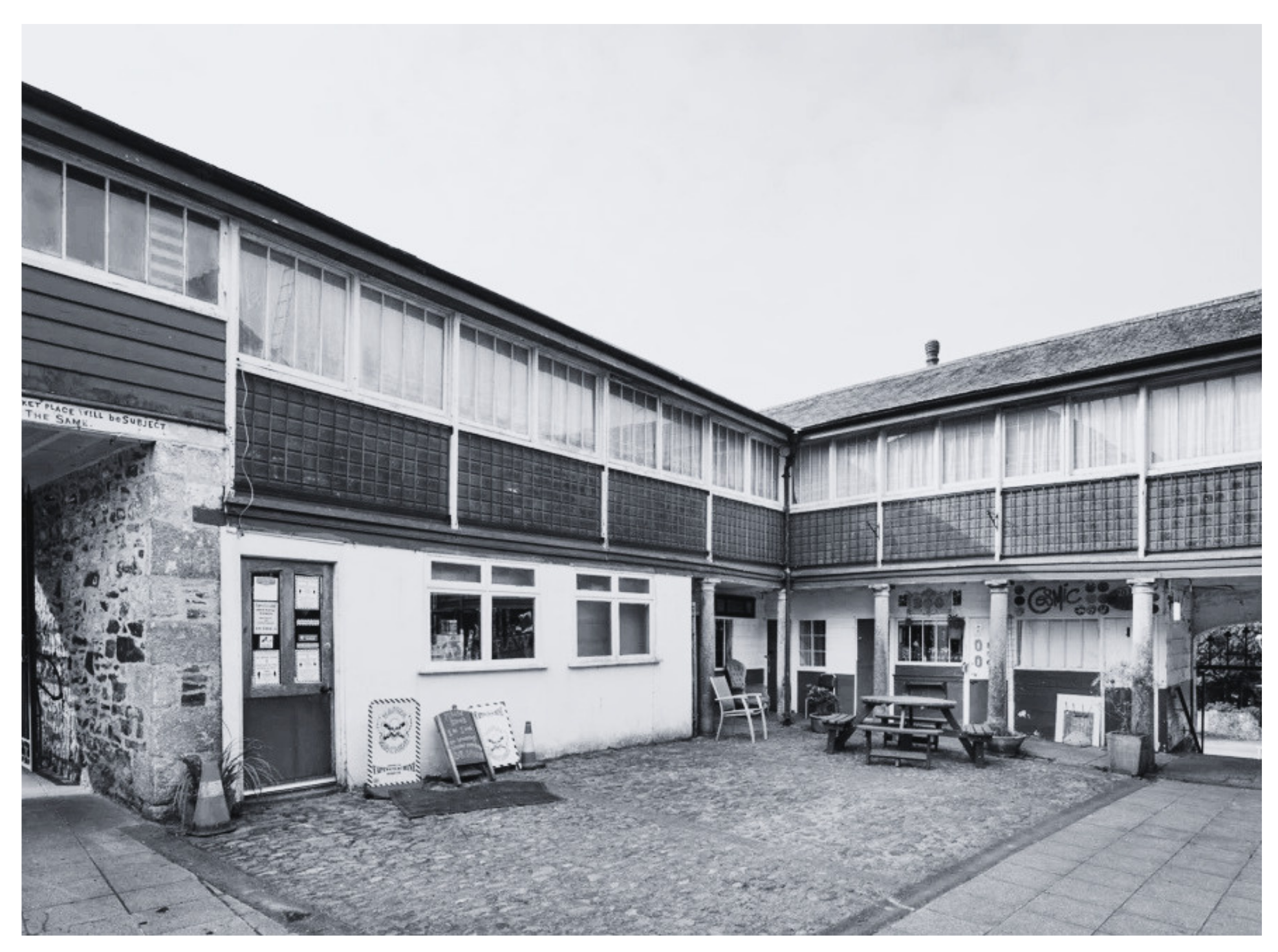 Black and white photograph of a building with large windows and a patio area with tables, chairs, and decorative signs outside.