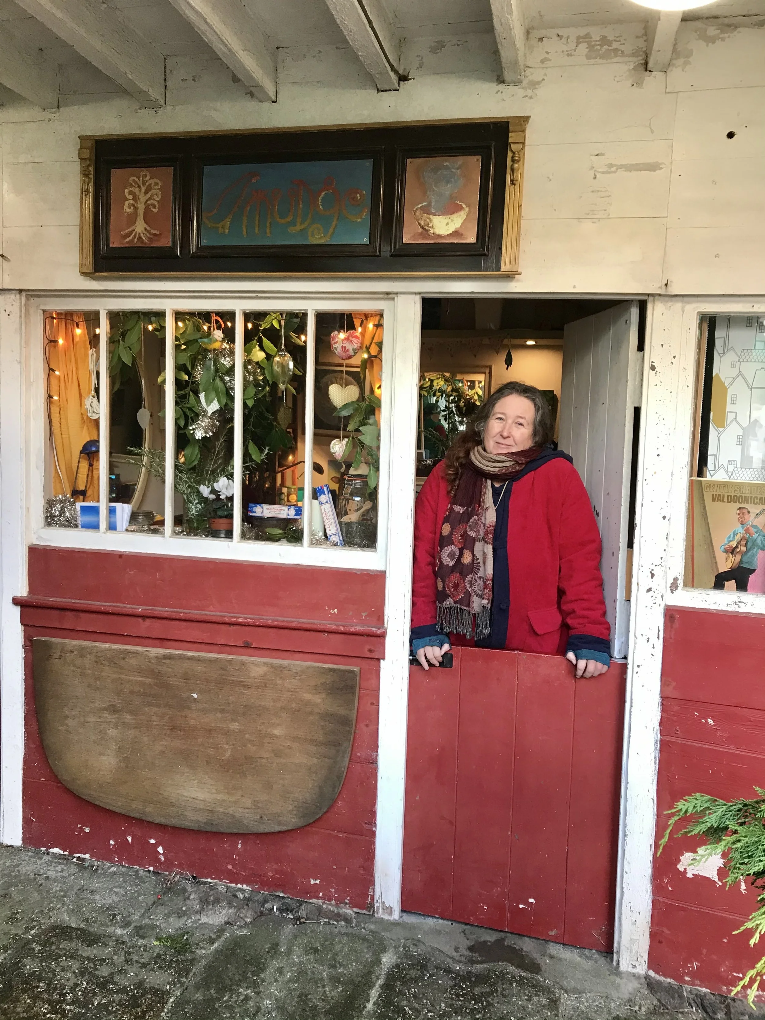 A woman with long brown hair wearing a red jacket and scarf standing behind a rustic red and white wooden booth or window with a waffle design on it. Inside the booth are plants, hanging ornaments, and lights.