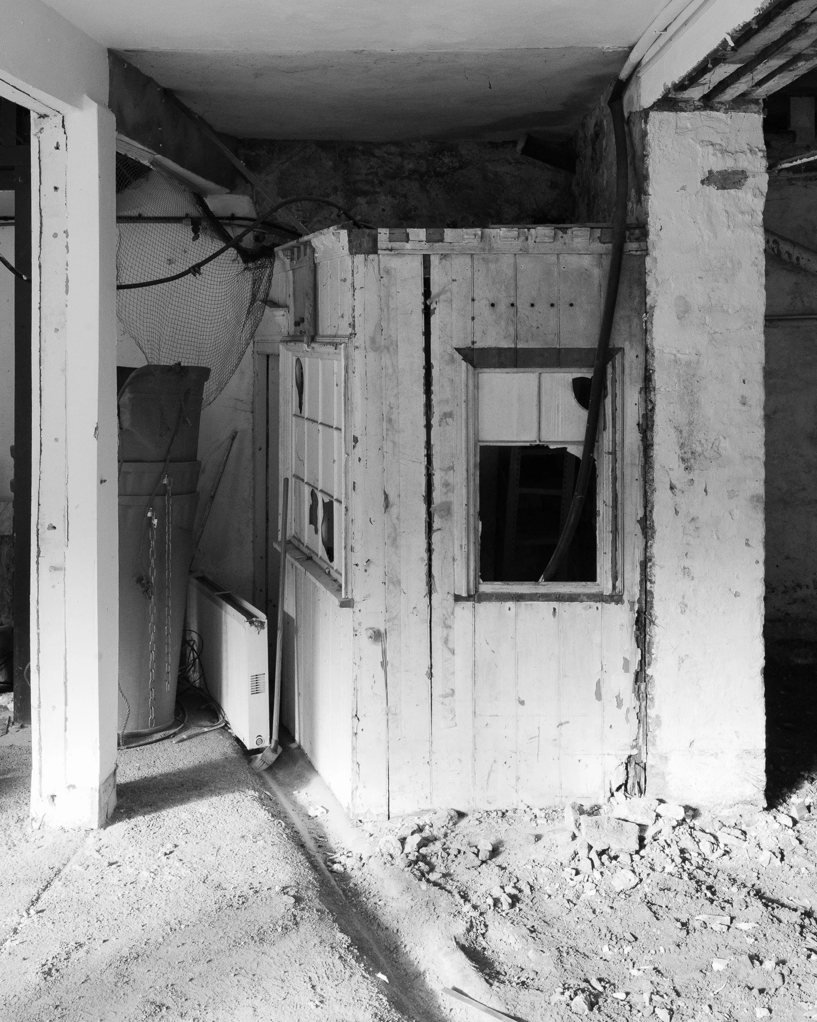 Construction site with debris, dirt, and rubble in a partially demolished room. An old wooden partition with a cut-out window, a loose cable, and a dust-covered vacuum cleaner are visible.
