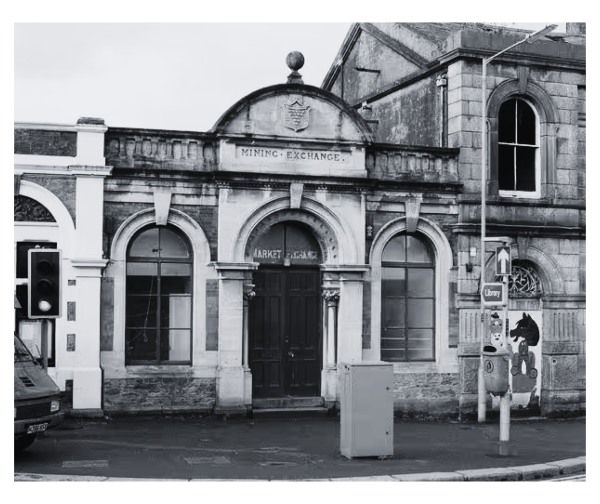 Black and white photo of an old building with a sign that reads 'Mining Exchange'. There are arched windows, a decorative crest, and a lightning rod on the roof. A traffic light and part of a car are visible in the foreground.