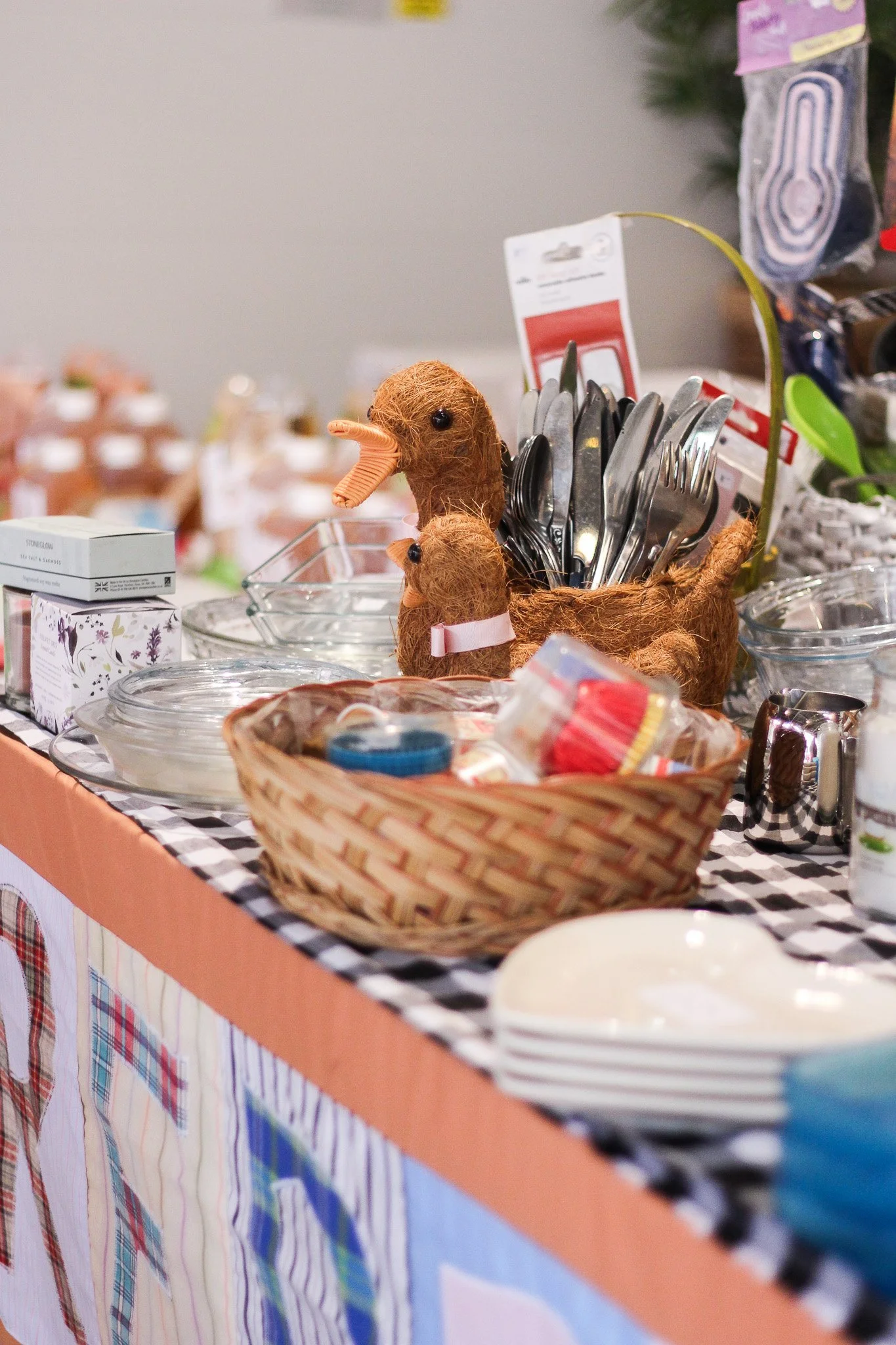 Table with various kitchen items including a woven basket, stacked plates, and cutlery, with decorative plaid fabric on the tablecloth.