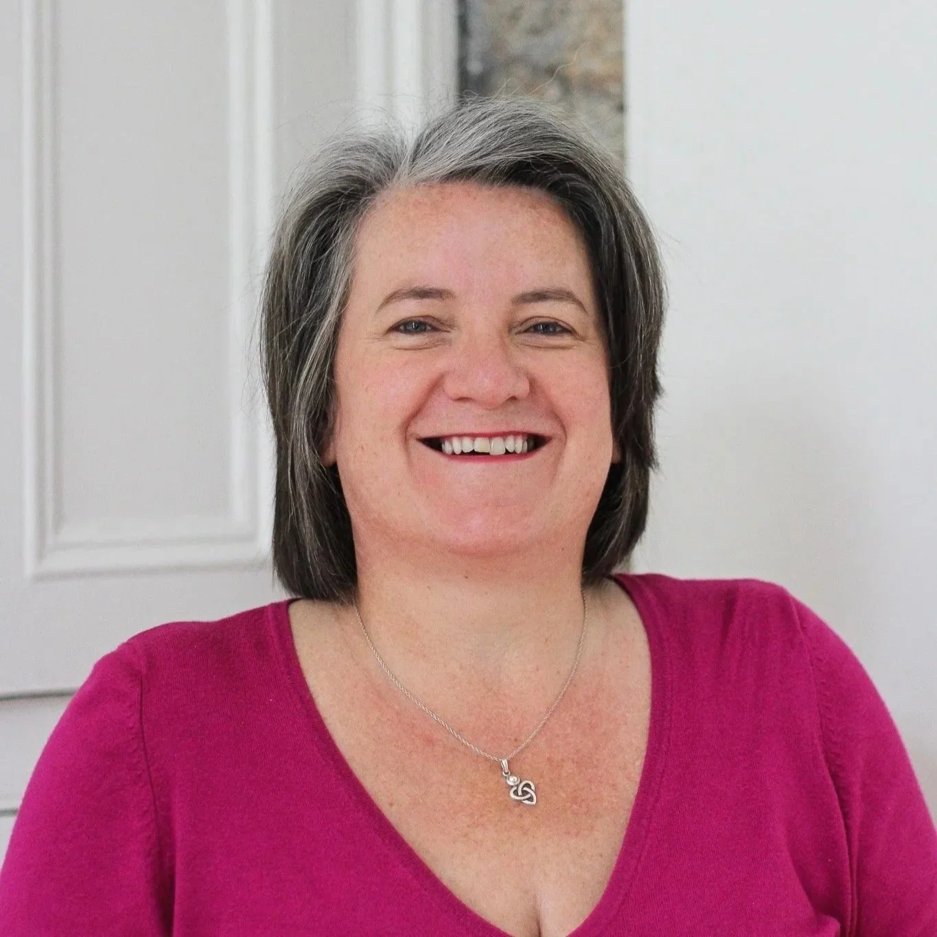 A woman with short gray hair, smiling, wearing a pink top and a silver necklace with a heart-shaped pendant, standing in front of a white wall.