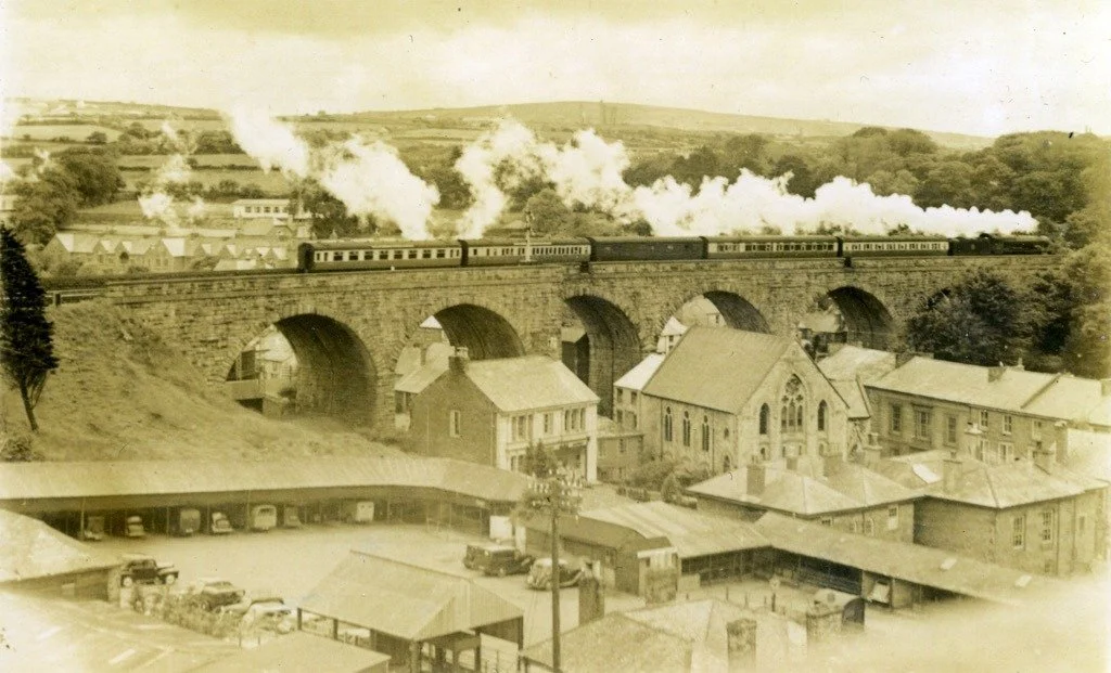 An old sepia-tone photograph of a steam train crossing a stone bridge over a town, with buildings and hills in the background.