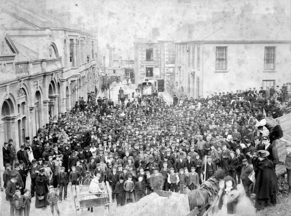 Crowd of people gathered outside buildings on a city street in the early 20th century, with some sitting on a ledge overlooking the crowd.
