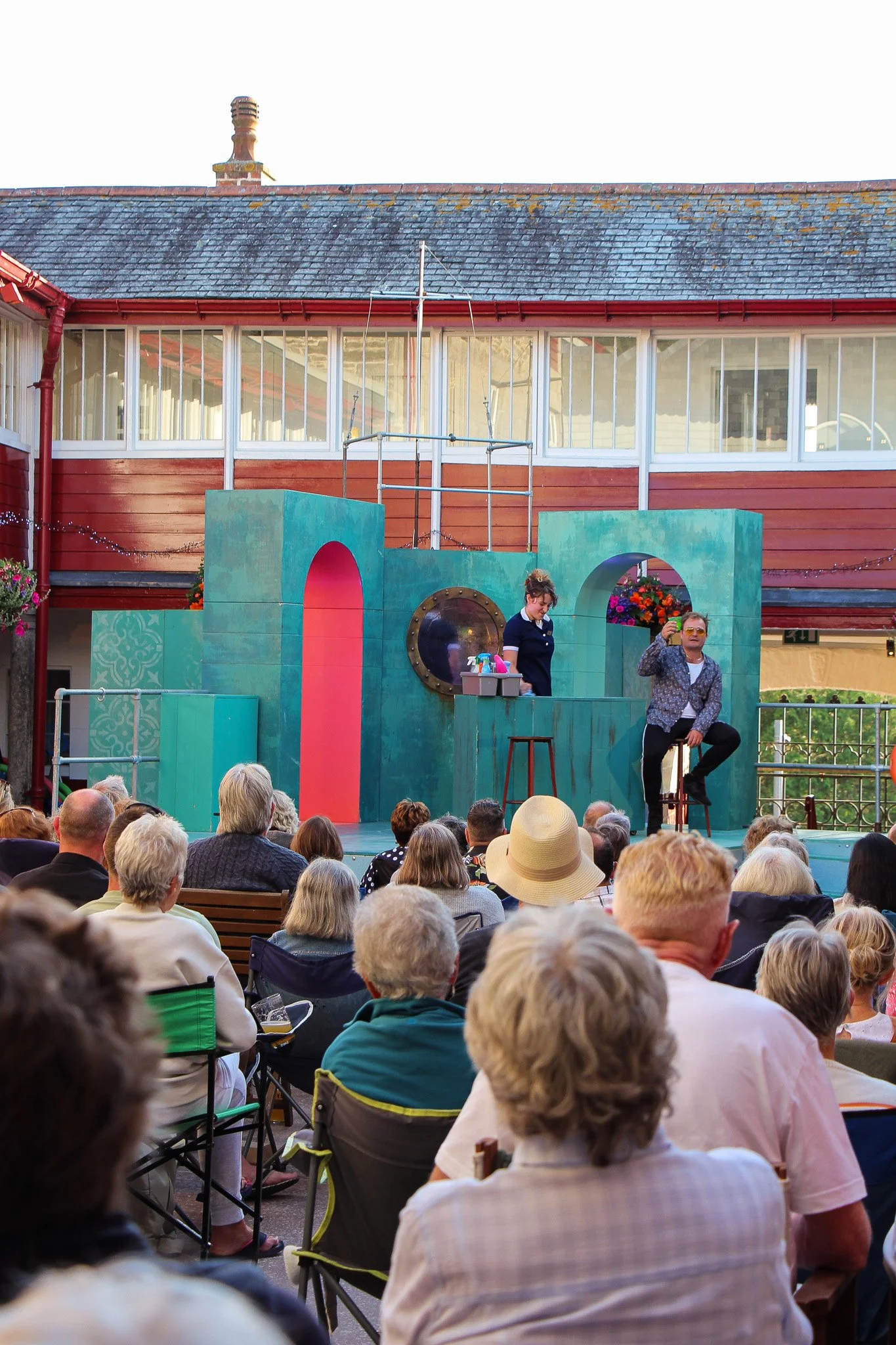 Audience watching an outdoor theatrical performance featuring two actors and a colorful, decorative set design in front of a building with large windows and a sloped roof.