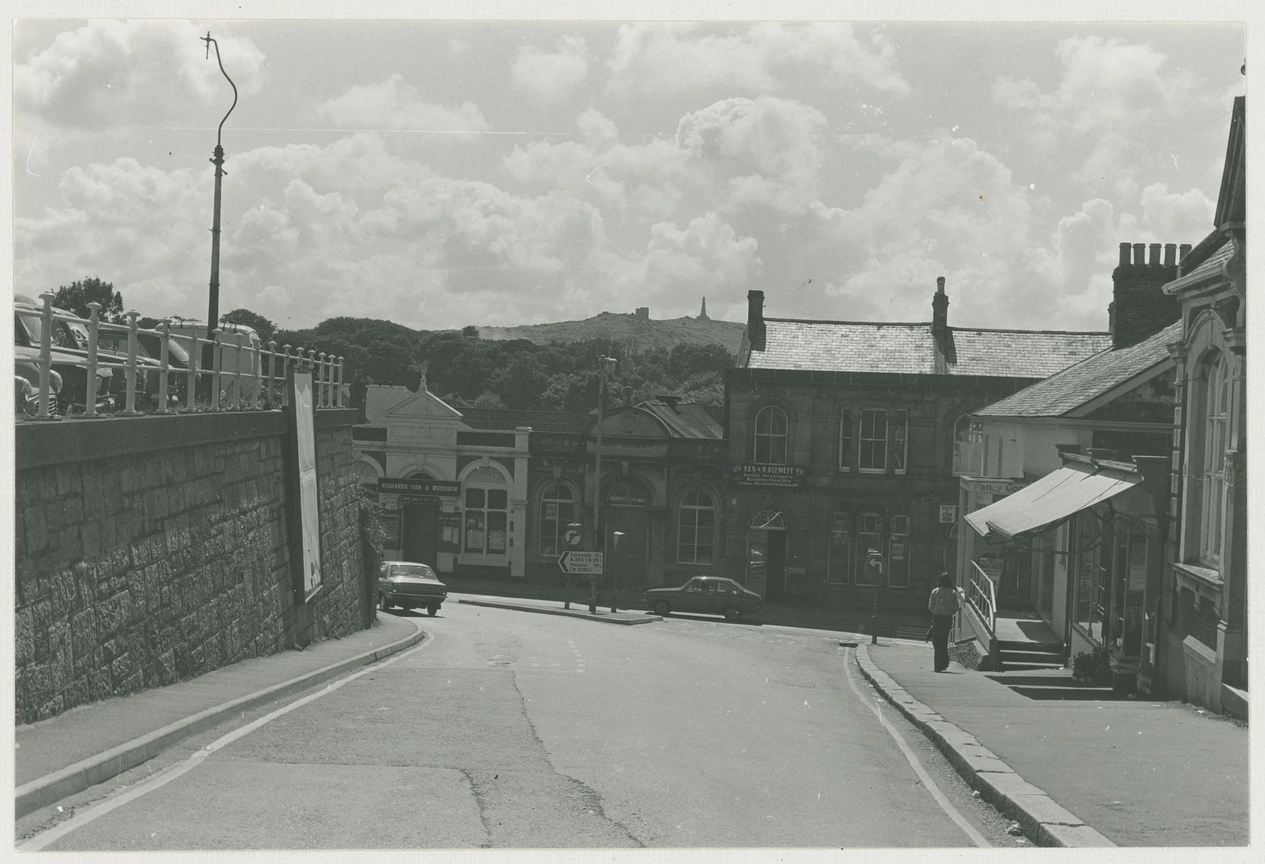 Black and white photo of a small town street with commercial buildings, parked cars, and a hill with a monument on top in the background. Overcast clouds in the sky.