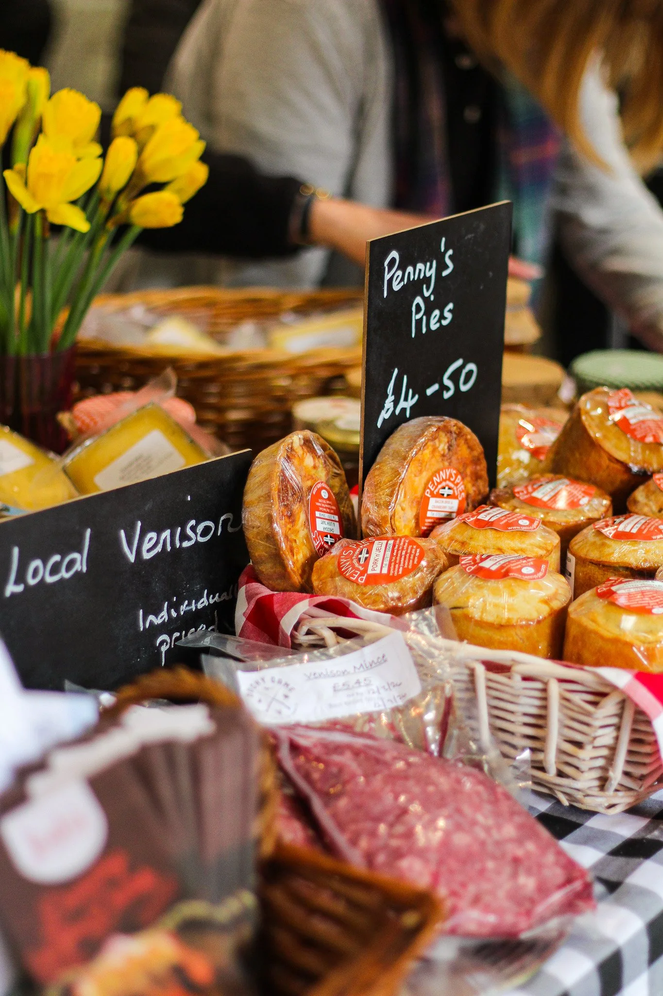 Display of various local venison and pork pies on a market stall, with a black chalkboard sign reading 'Penny's Pies' and the price of £4.50, decorated with yellow flowers and other food items in the background.