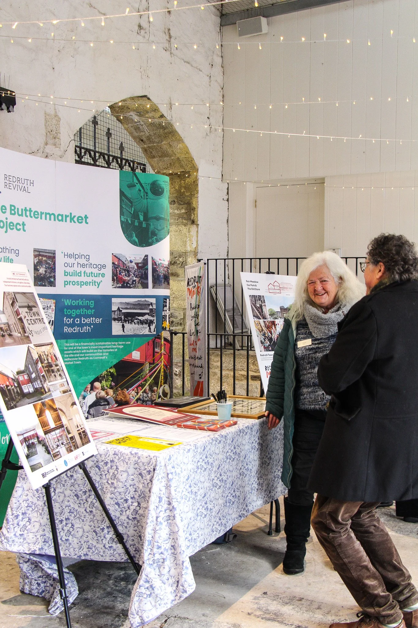 People in conversation at a community event or exhibition, with informational posters and displays about a project named 'Redruth Revival' and 'The Buttermilk Project'.