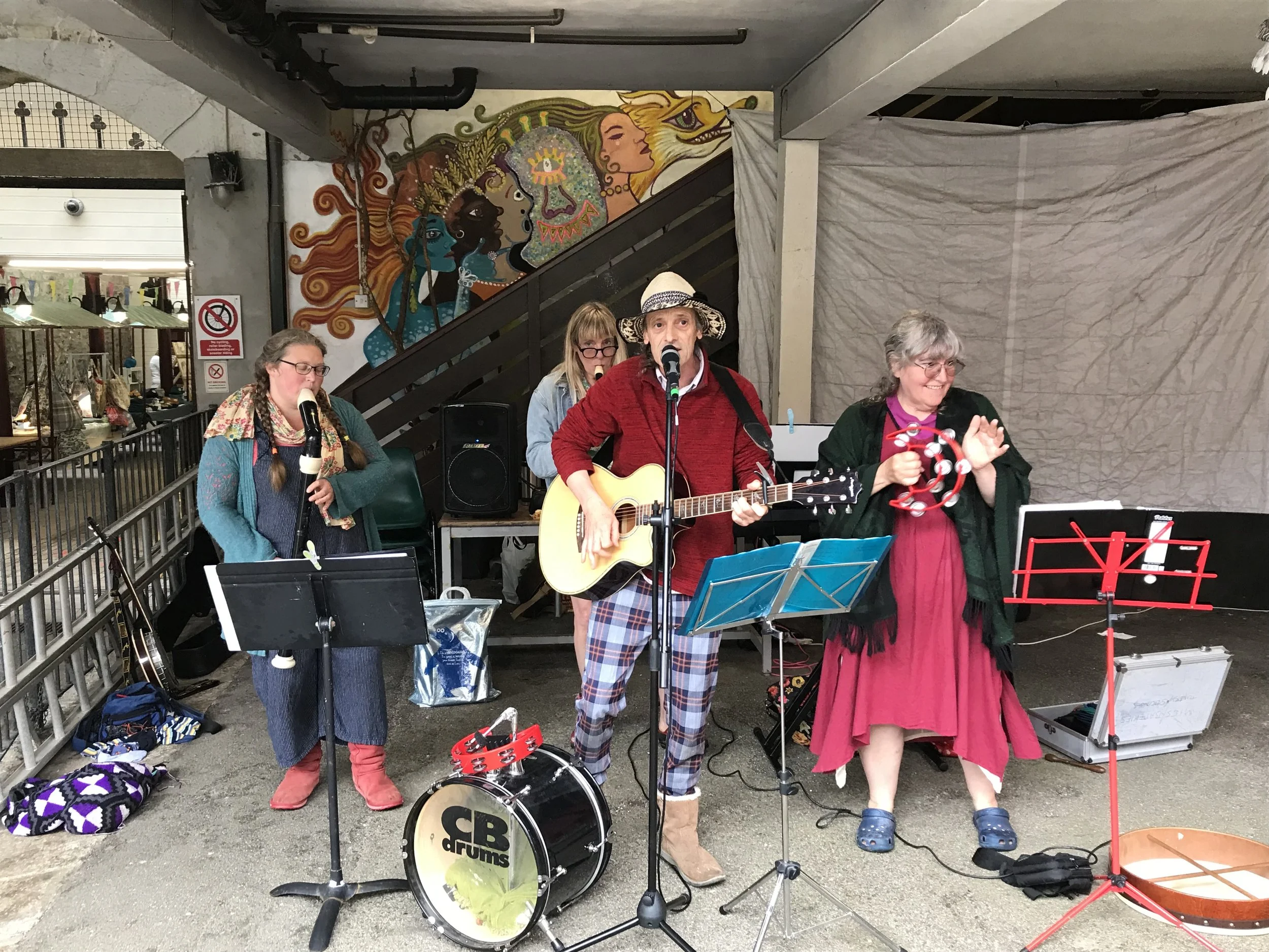 A group of four street musicians performing with instruments in an outdoor area. There is a colorful mural of stylized women's faces with flowing hair in the background. The musicians are singing and playing guitar, drums, and tambourine, with music stands and equipment around them.