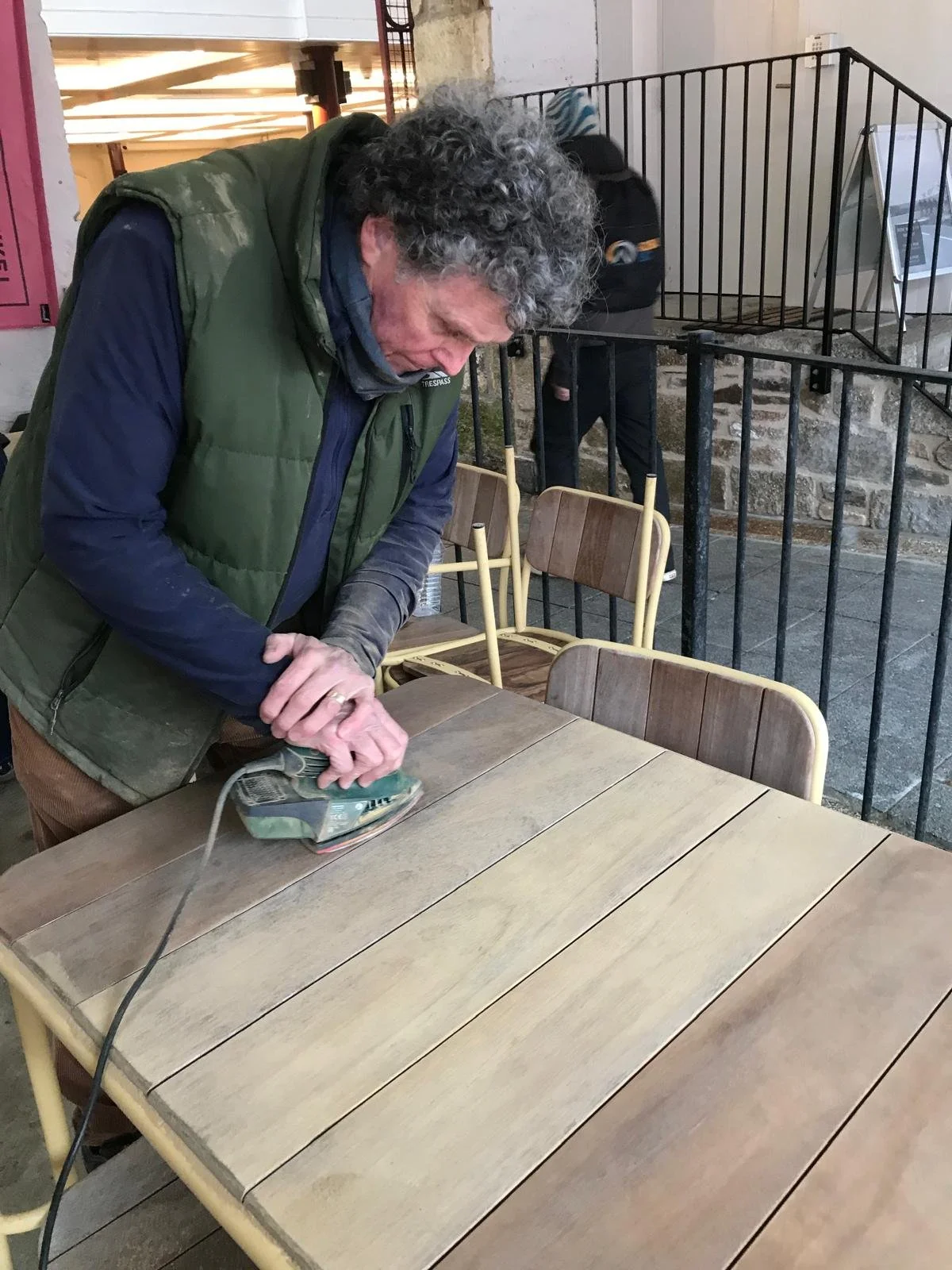 A man with curly gray hair is using an electric sander on a wooden table in a workshop or cafe setting.