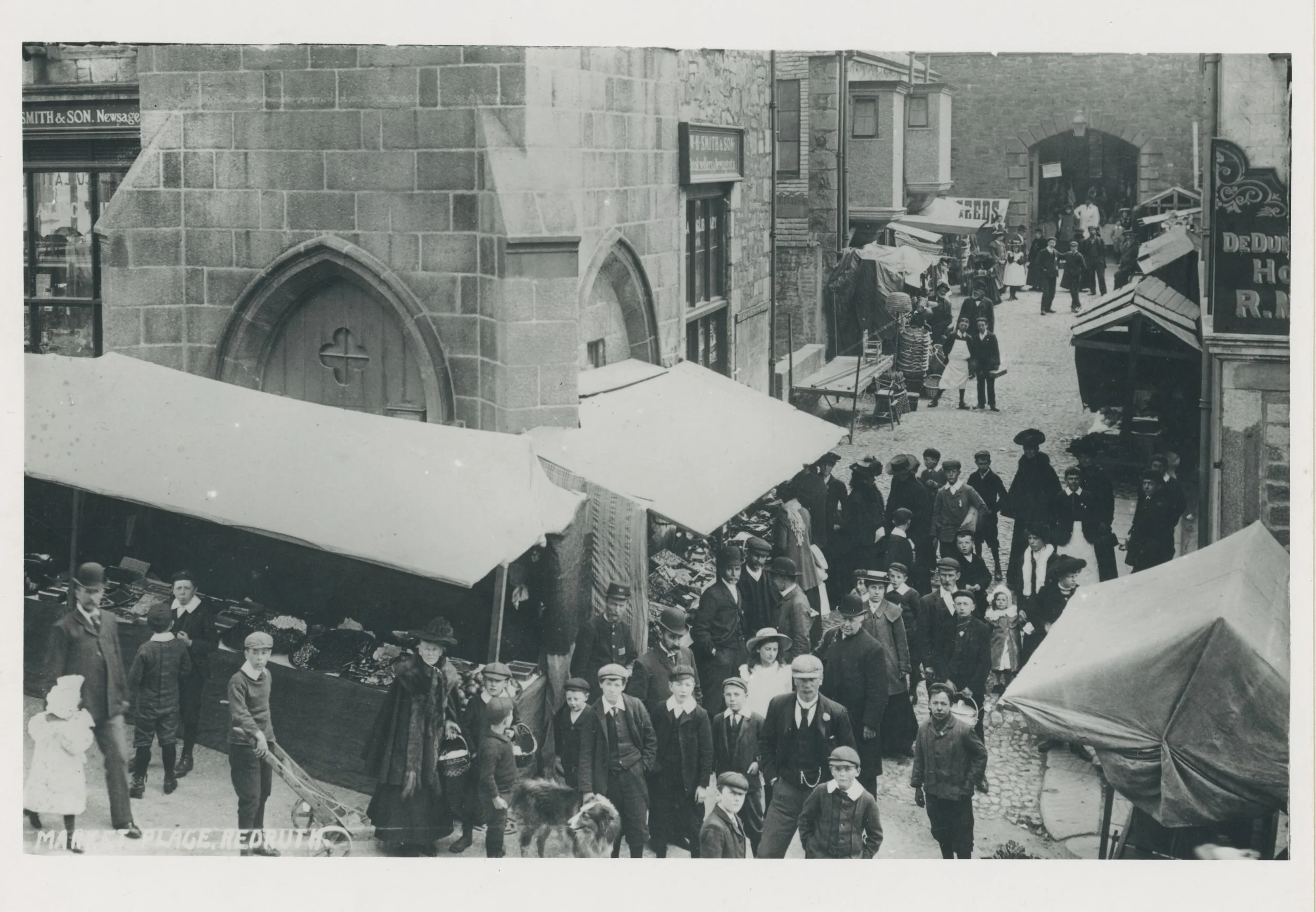 Historical black and white photograph of a crowded street market with people shopping and walking, old buildings, tents, and signs, including one that reads 'Smith & Son.'