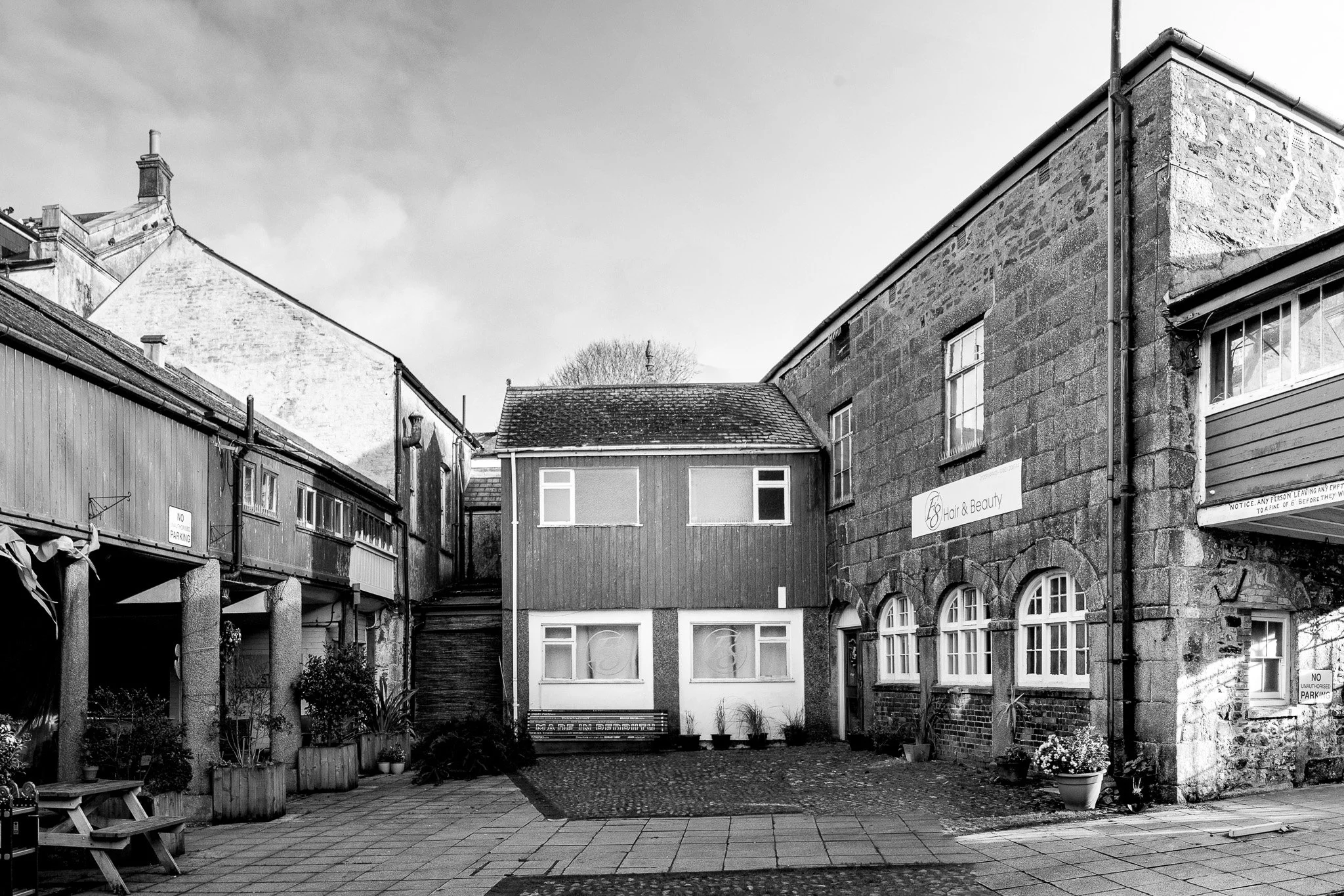 Black and white photo of a small urban courtyard surrounded by old stone and wood buildings with windows, potted plants, benches, and signs, including a 'No Parking' sign and a sign for a hair and beauty salon.