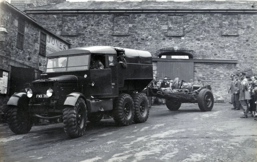 An old black and white photo of a vintage military truck with a flatbed trailer attached, parked in front of a stone building with a crowd of people nearby.