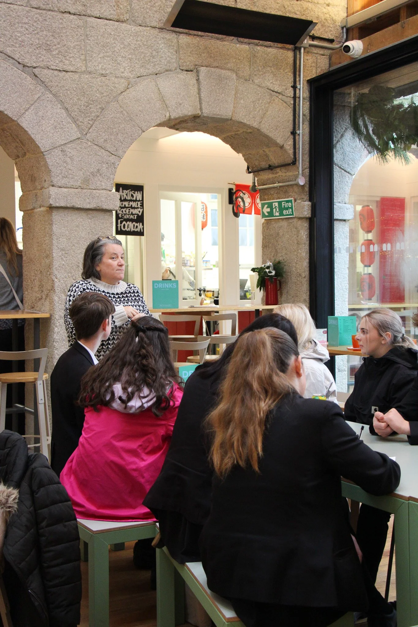 People seated at tables in a restaurant or café with stone arches, some standing and others sitting. A woman in a black and white patterned sweater is standing, and a woman in a pink jacket is sitting with her back to the camera. A small chalkboard menu, a fire exit sign, and a security camera are visible on the stone wall.