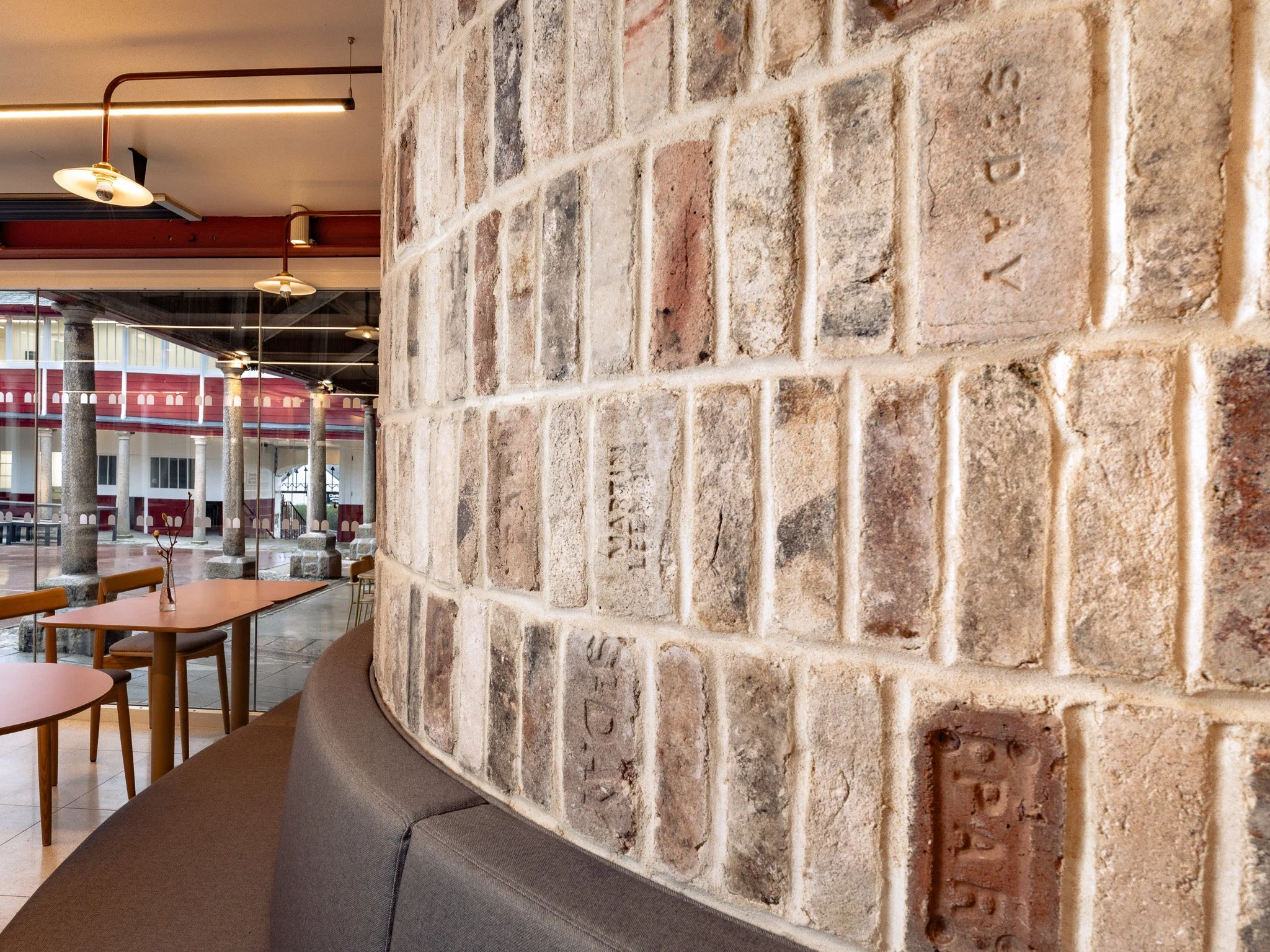 Interior of a modern cafe or restaurant with brick walls, wooden tables, and chairs. Large windows and exposed beams are visible, along with hanging lights.