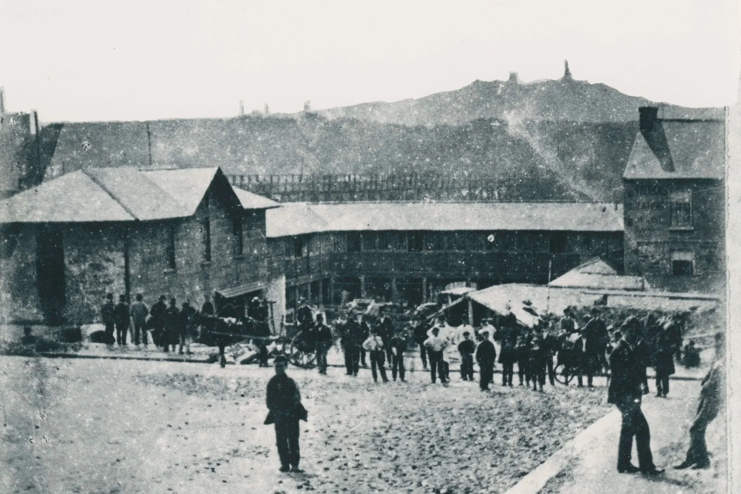 Black and white historical photograph of a city square with people, horses, and wagons. In the background, there is a large building with a tower or steeple and a view of a mountain with several structures or towers on top.
