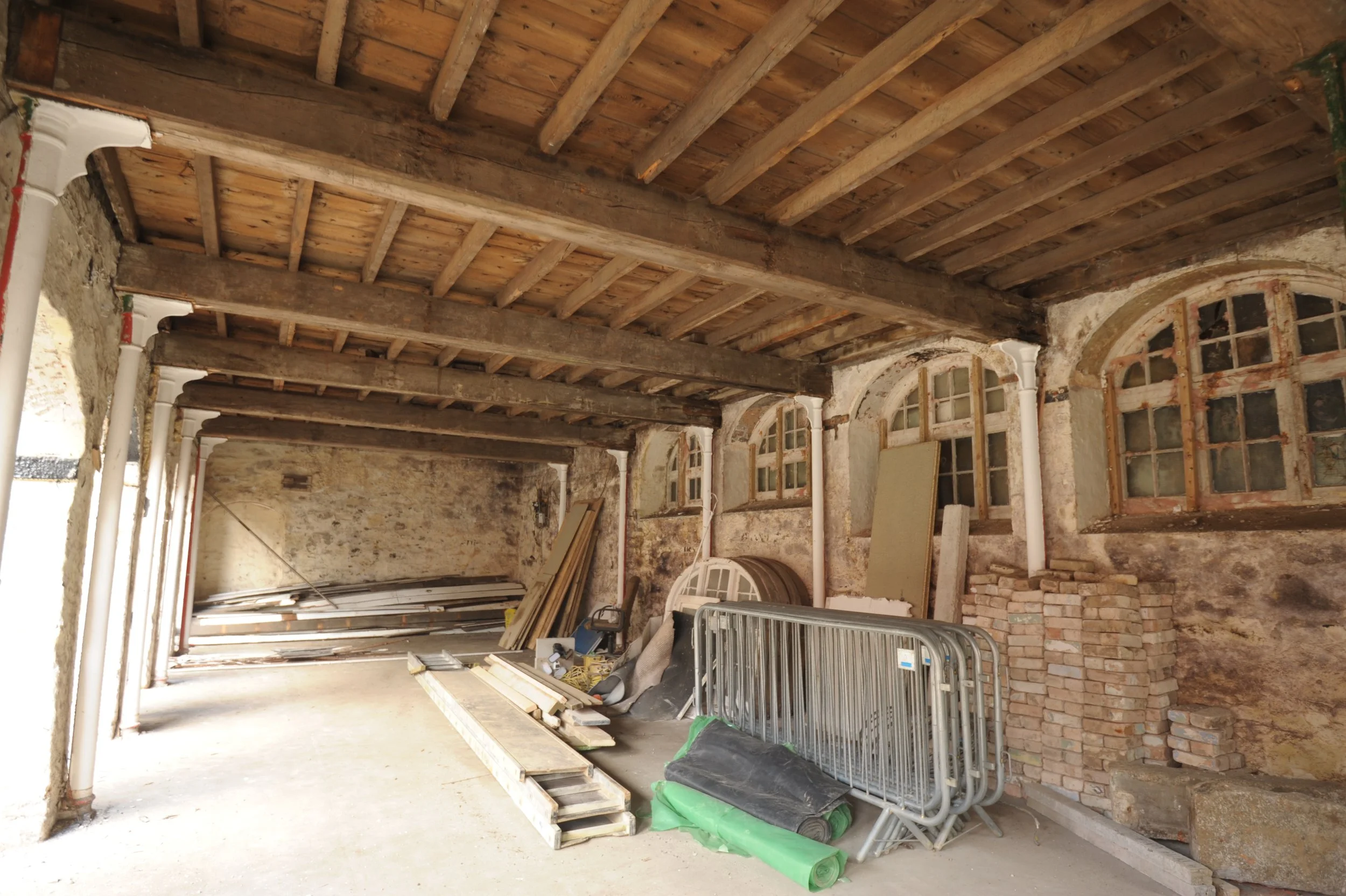 Construction site with exposed wooden beams on ceiling, brick walls, and construction materials including metal frames and rolled green insulation on the floor.