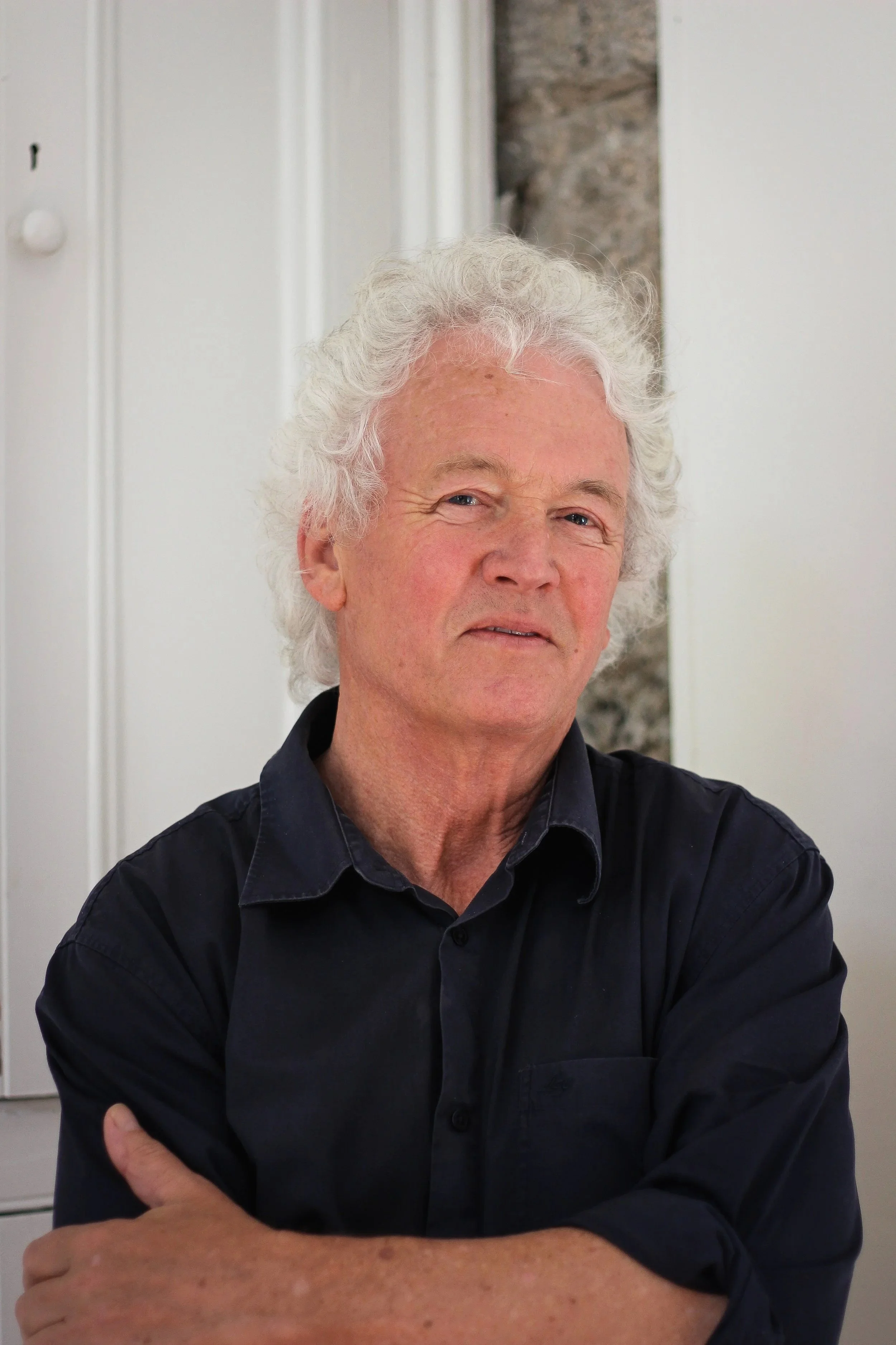 A man with curly gray hair and fair skin, wearing a black shirt, standing indoors near a wall with vertical white panels and a section of exposed brick behind him, with his arms crossed.