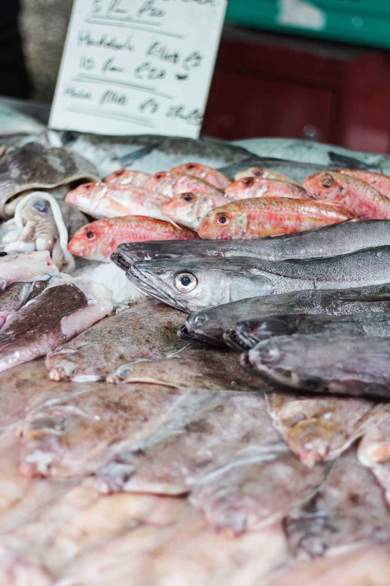 Fresh fish and seafood displayed on ice at a market stall, with a handwritten sign in the background.