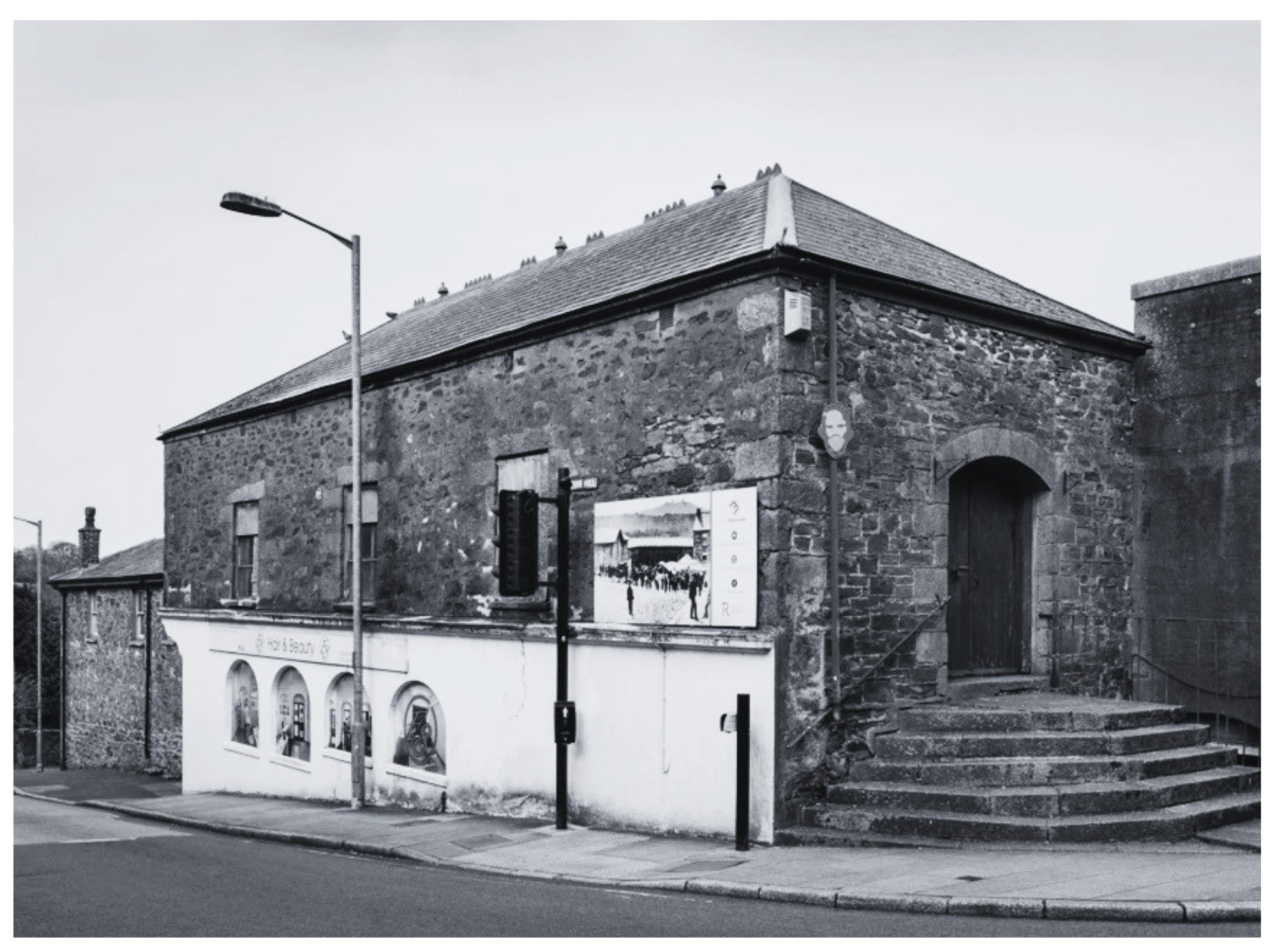 Black and white photo of an old brick building with a tiled roof, a street lamp, a traffic light, and a modern advertising poster on the wall.