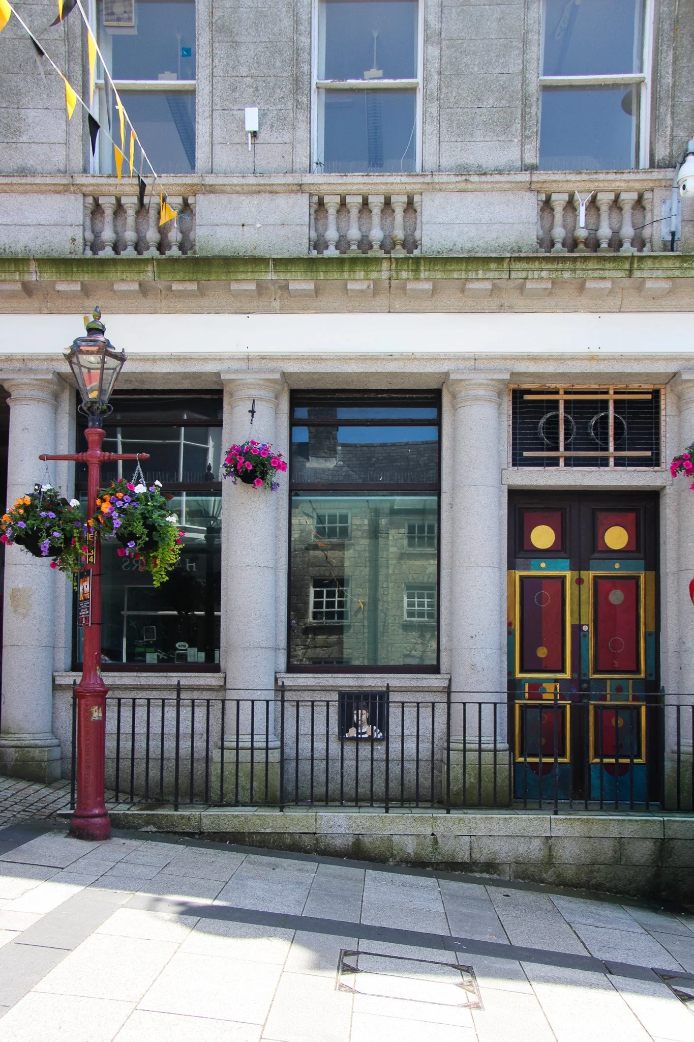 Front view of a building with large windows, stone columns, and a decorated door, surrounded by a black metal fence. There is a red lamppost with hanging flower baskets and a person sitting on the other side of the fence.