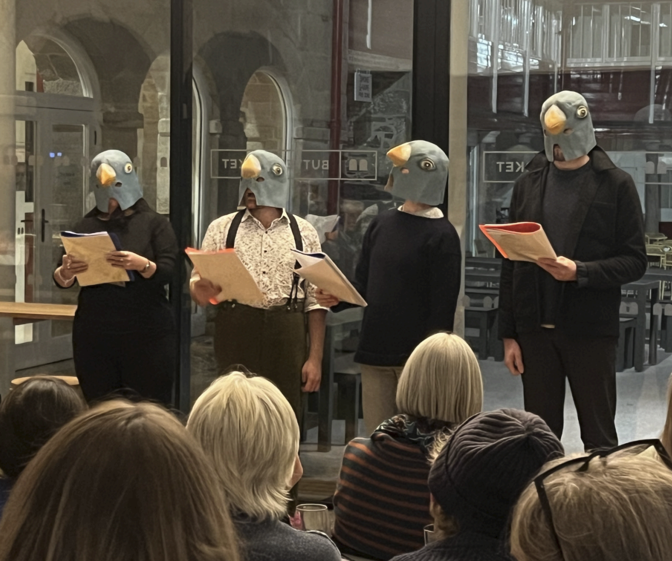 Four people wearing bird masks and holding papers stand in front of an audience inside a building.