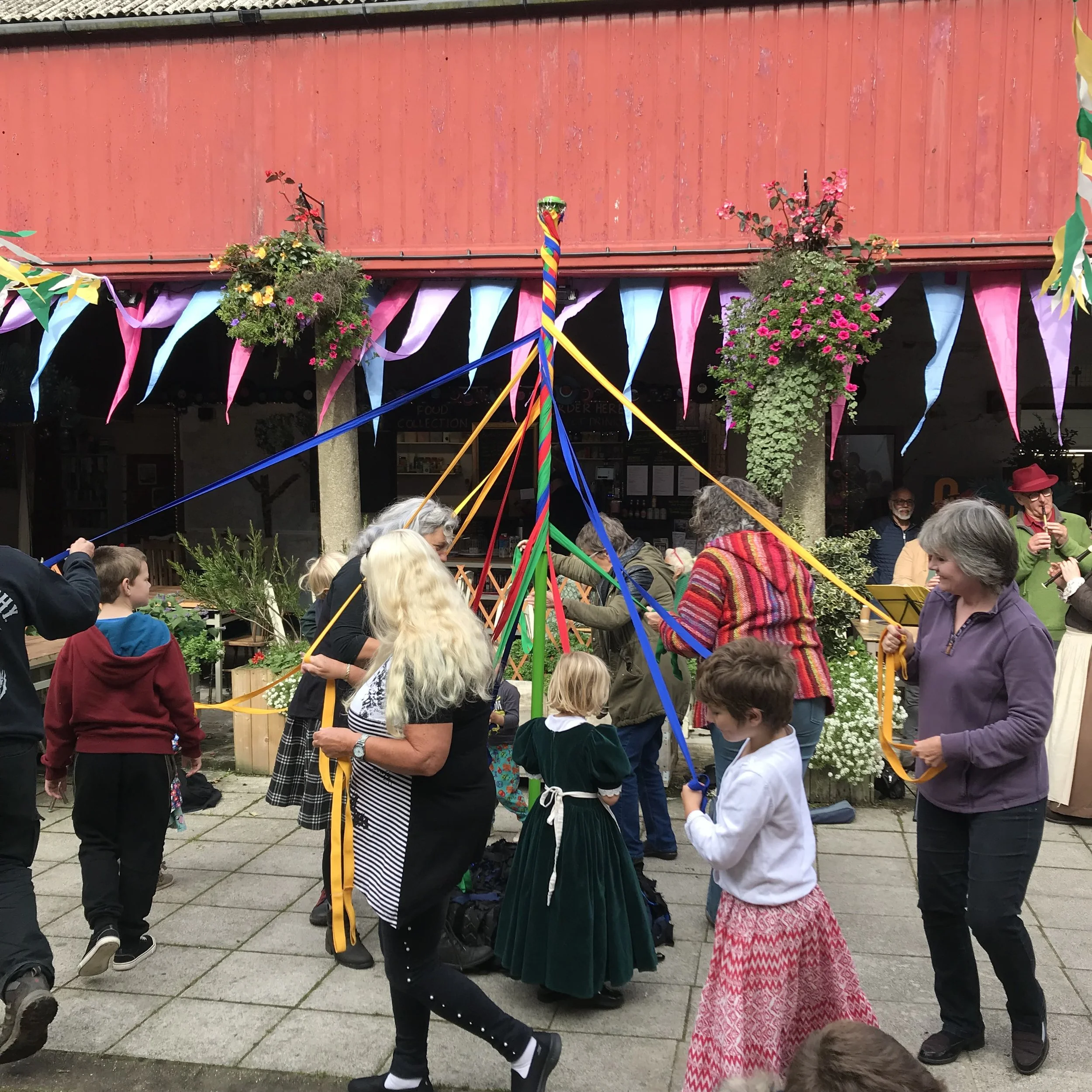 A group of people gather outdoors in front of a red building, participating in a celebration involving a maypole decorated with rainbow ribbons. The scene includes children and adults, some wearing colorful clothing, with flowers and bunting hanging across the area.
