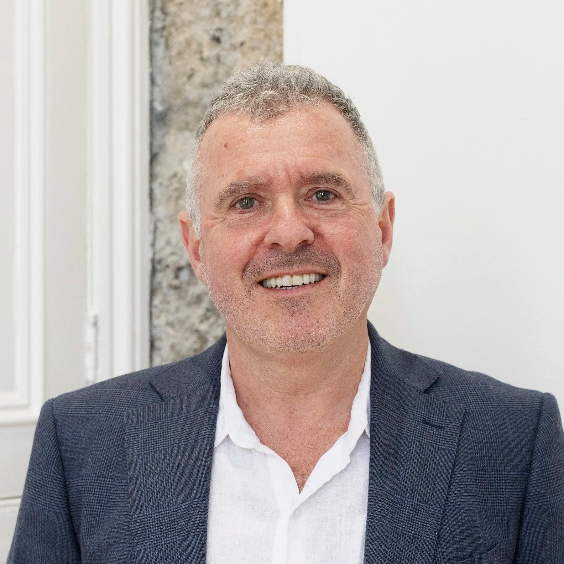 A man with gray hair, blue eyes, and a broad smile, wearing a white shirt and a dark plaid blazer, standing indoors with a partially visible white wall and stone wall in the background.