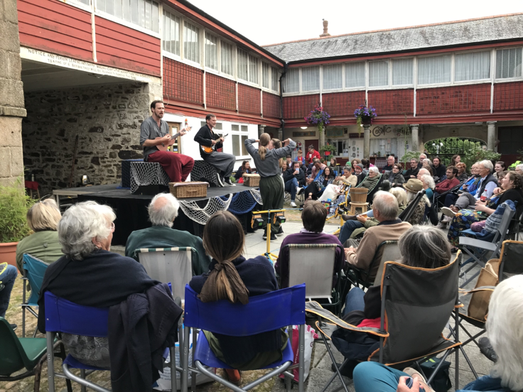 A group of people is watching a live outdoor performance with two musicians, one playing guitar and the other playing a melodica, in a courtyard surrounded by red and stone buildings. An audience of various ages is seated in chairs, listening to the music.