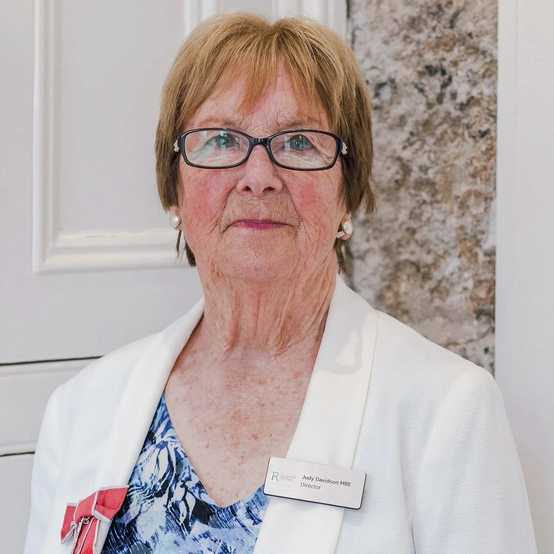 Close-up of an elderly woman with short reddish hair, glasses, and a serious expression. She is wearing a white jacket, a patterned blouse, and pearl earrings. The woman has a name tag that reads Judy Davidson MBE, Director. The background includes a white wall and a marble surface.