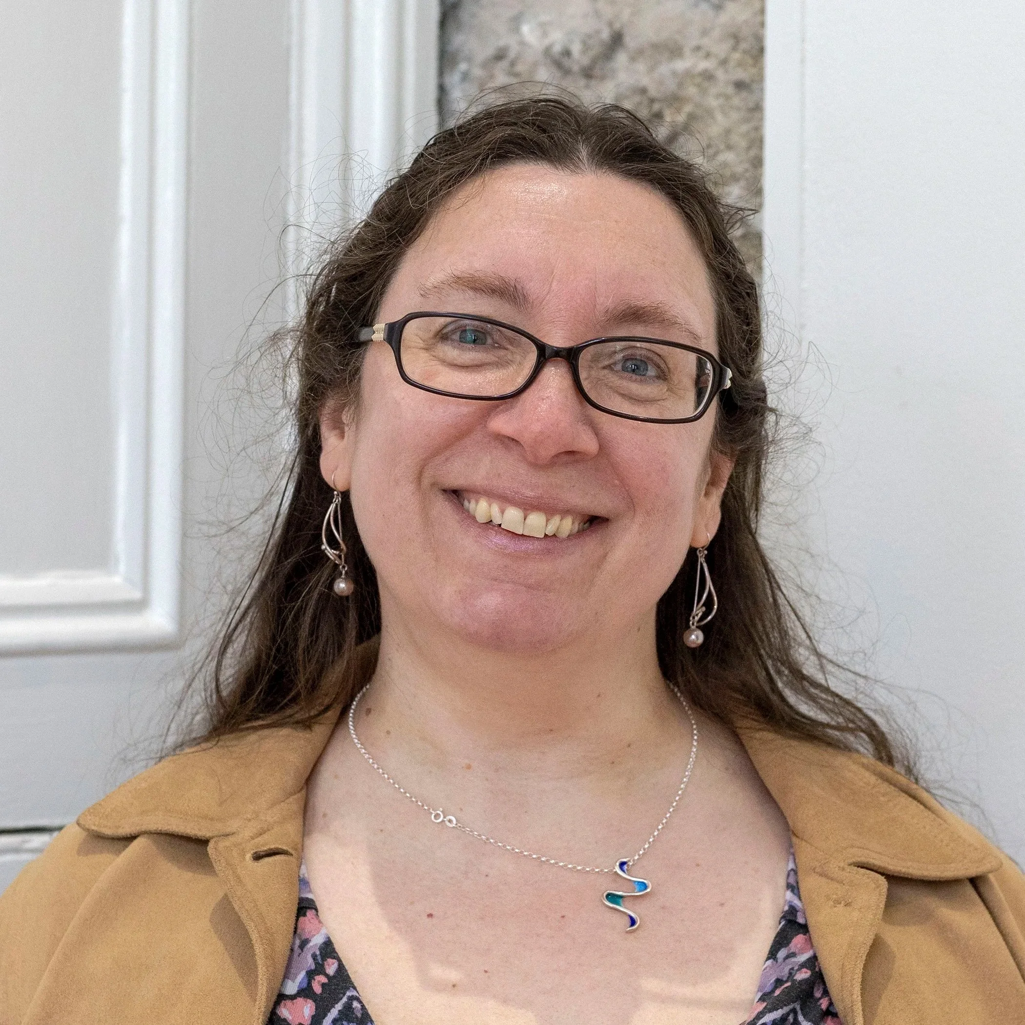 A smiling woman with glasses and earrings wearing a beige blazer and a necklace with a colorful wave pendant, standing in front of a neutral background.