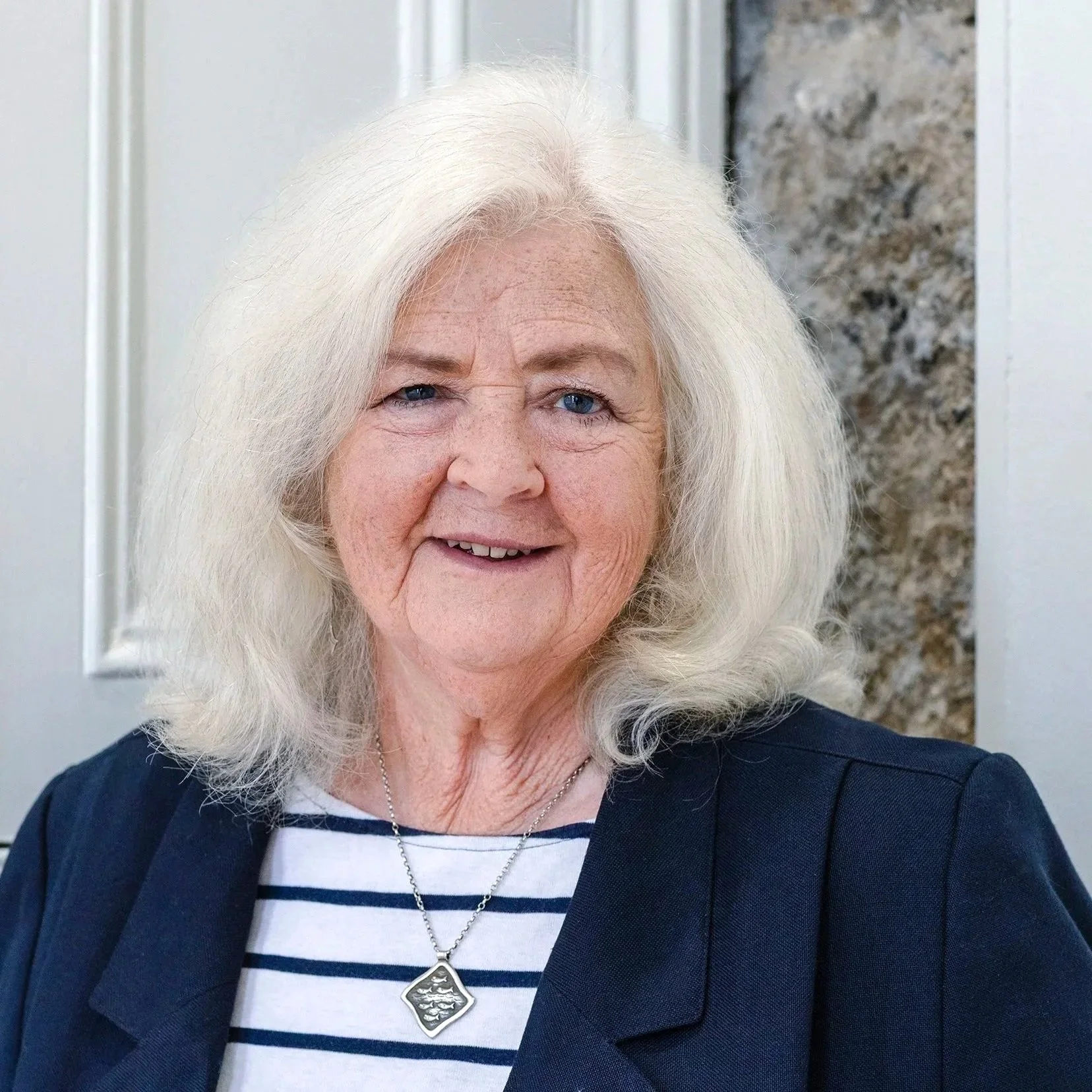 Close-up of a woman with white hair, blue eyes, smiling, wearing a striped shirt, dark blazer, and a silver necklace with a pendant.