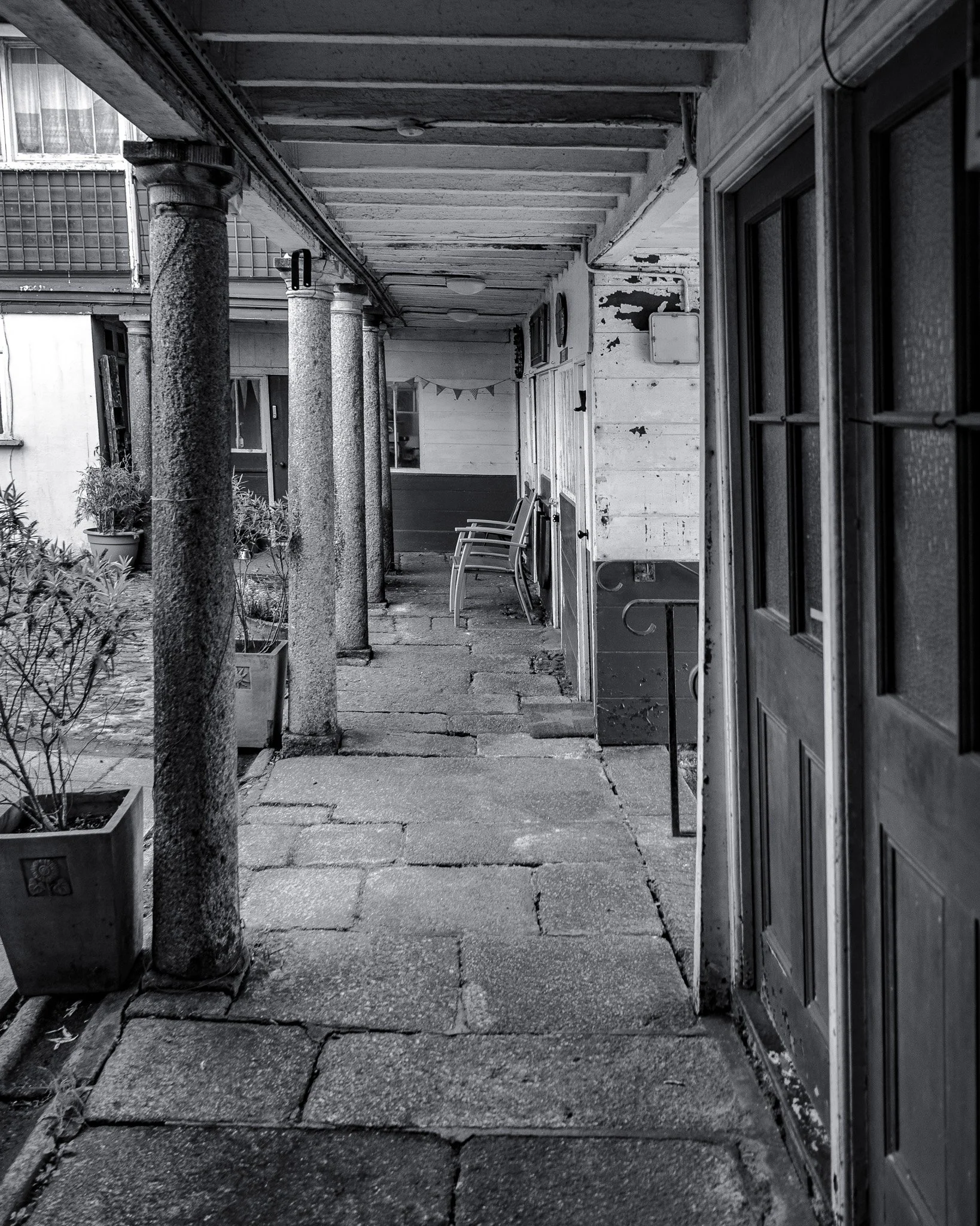 A black and white photo of a porch with concrete floor and ceiling, multiple stone columns supporting the roof, a small potted plant on the left, a few chairs along the wall, and a window and door on the right side.
