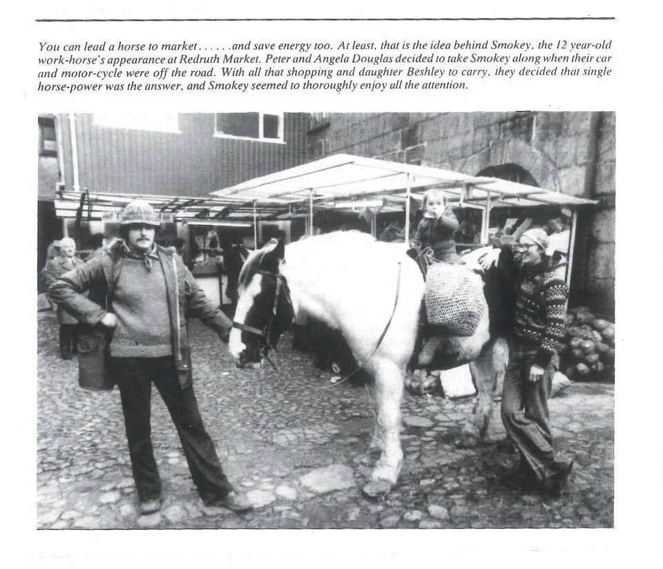 Black-and-white photo of two men standing next to a white horse with a child riding on its back, set in an outdoor market scene with a building and market stalls in the background.