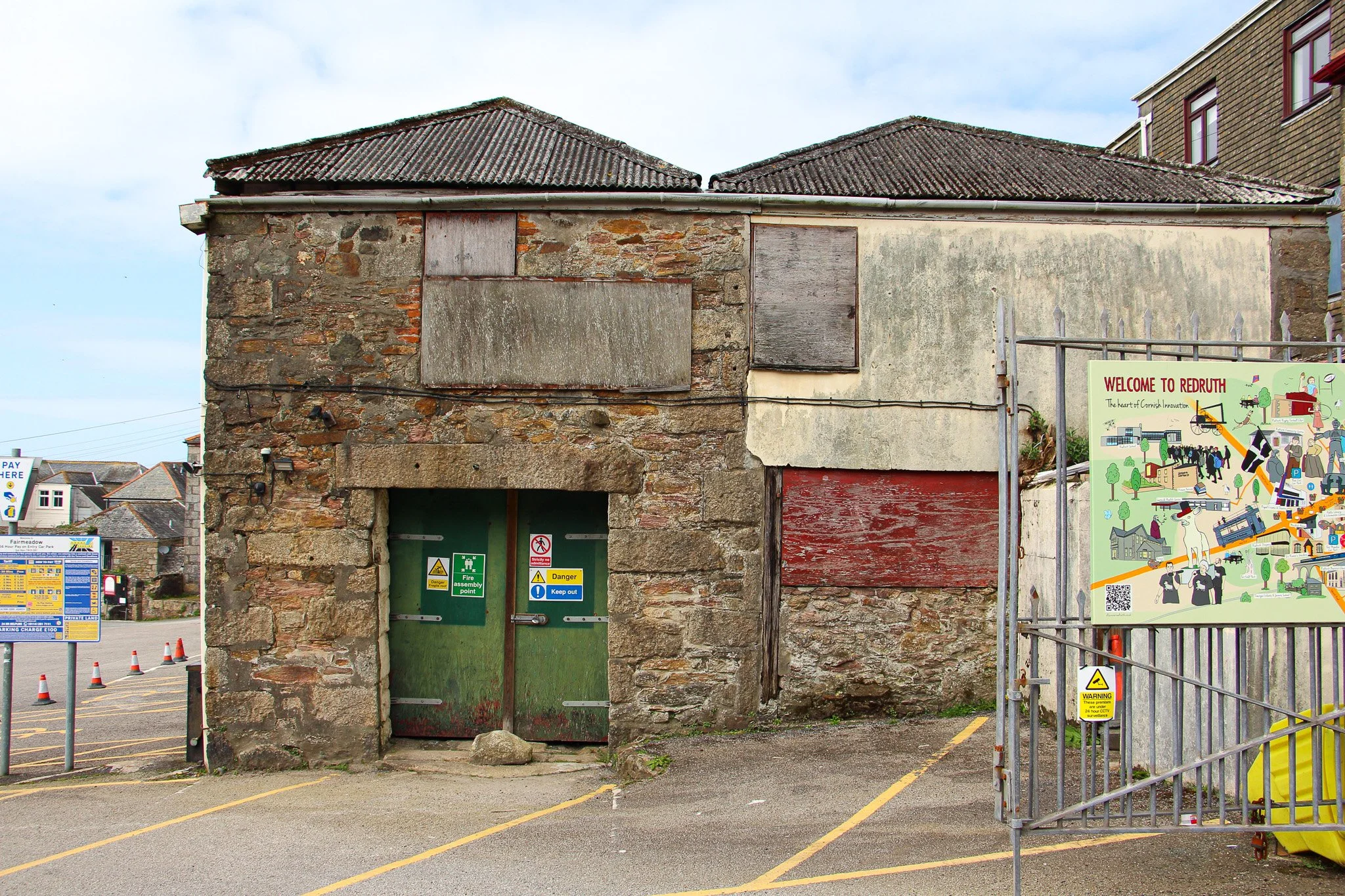 An old stone building with boarded-up windows and a green door, situated in a parking lot with orange traffic cones and various warning signs, next to a colorful informational sign about Redruth, Cornwall.