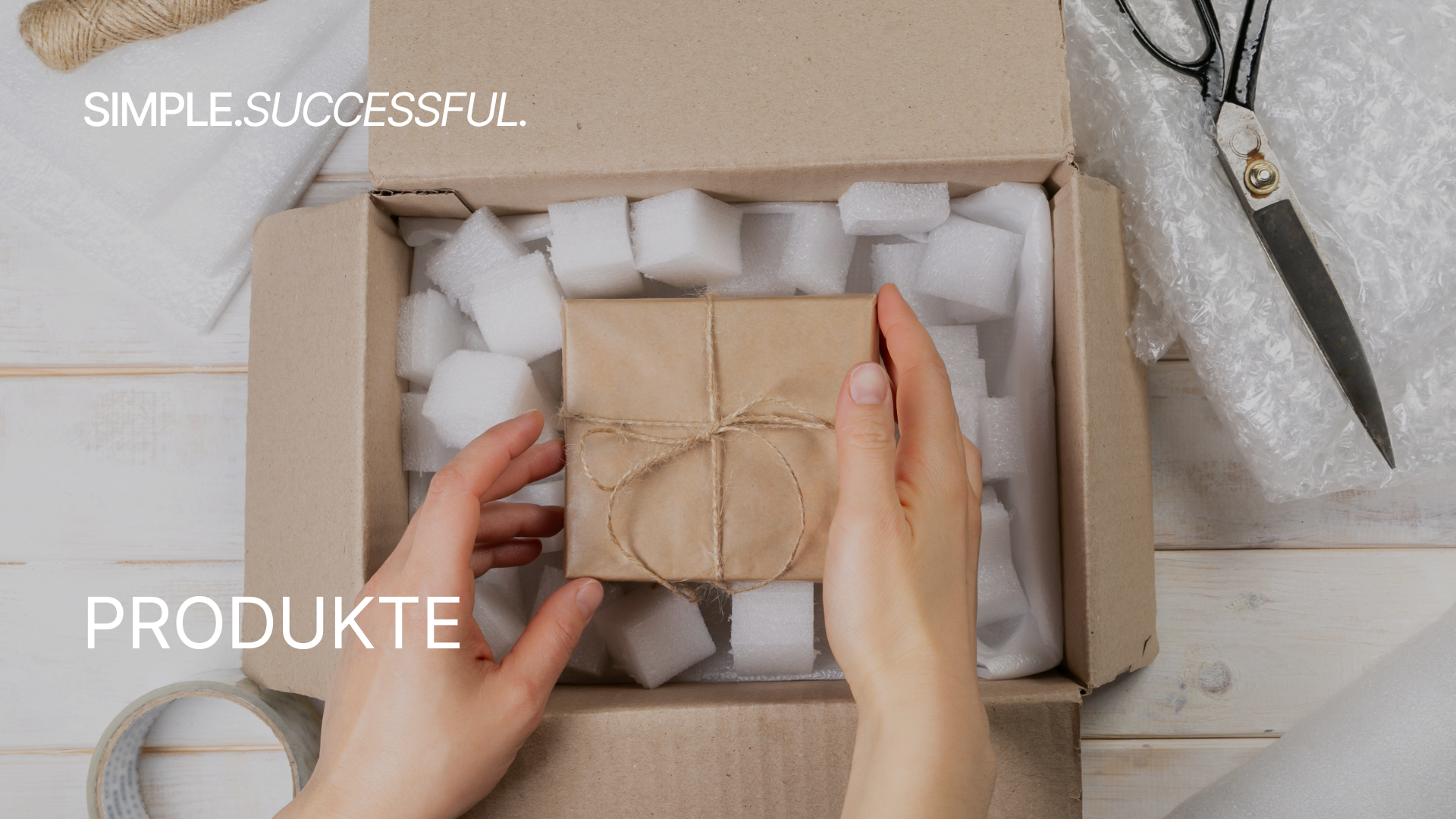 Hand hält ein Geschenk in braunem Papier mit Juteschnur vor ein Paket mit Styroporeinlage, umgeben von Verpackungsmaterialien, Schneideschere, Klebeband und Luftpolsterfolie auf einem weißen Holztisch.