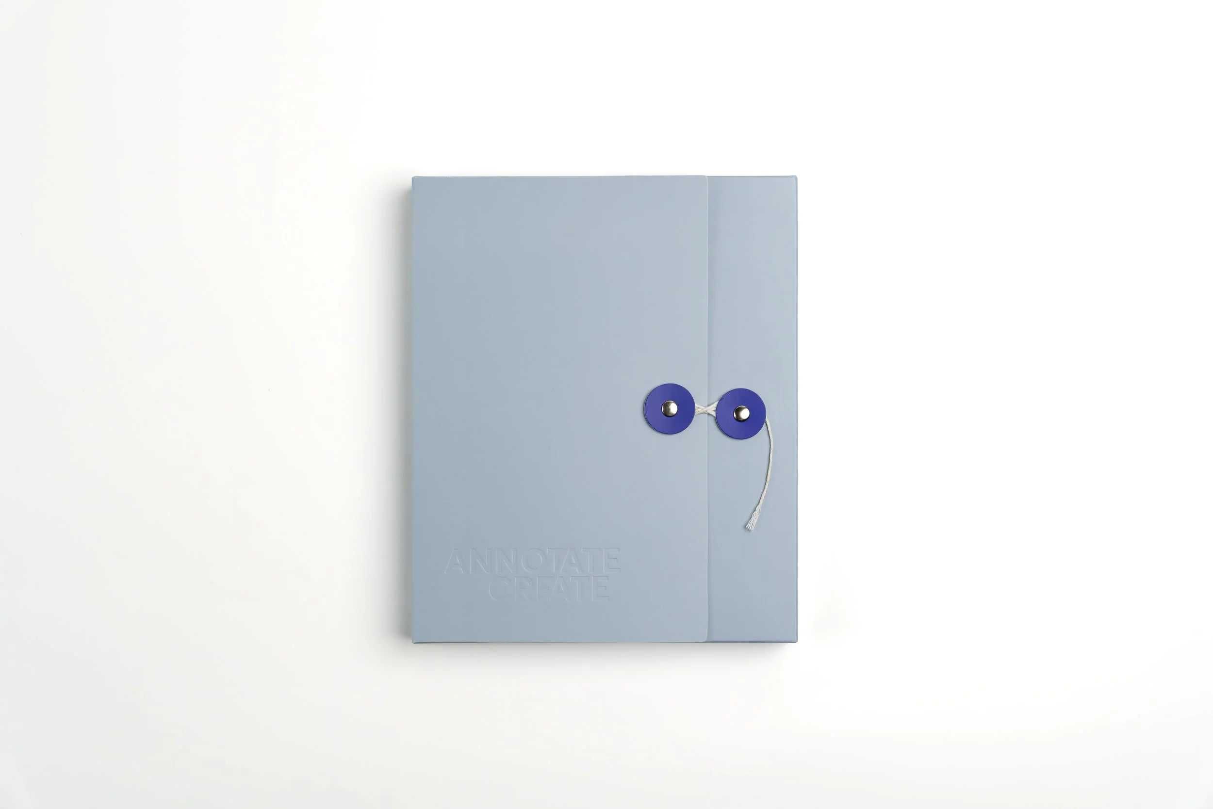 A light blue journal or planner with an attached white string, sitting against a white wall. The journal has the words 'annotate' and 'create' embossed near the bottom.