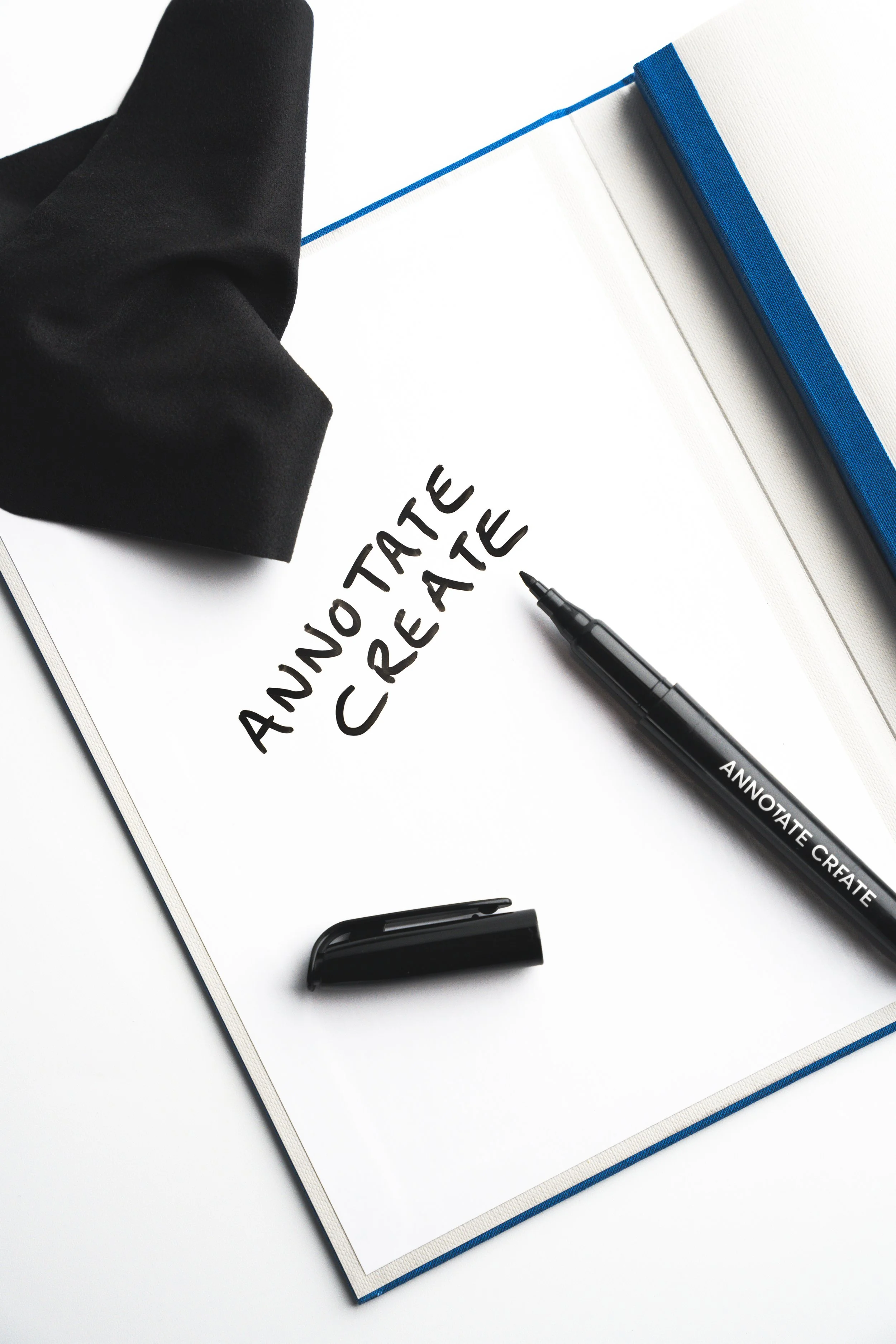 A white notepad with the words 'ADVOCATE CREATE' handwritten in black marker, black marker pen, a black highlighter, a black cloth, and a blue-edged white folder on a white surface.