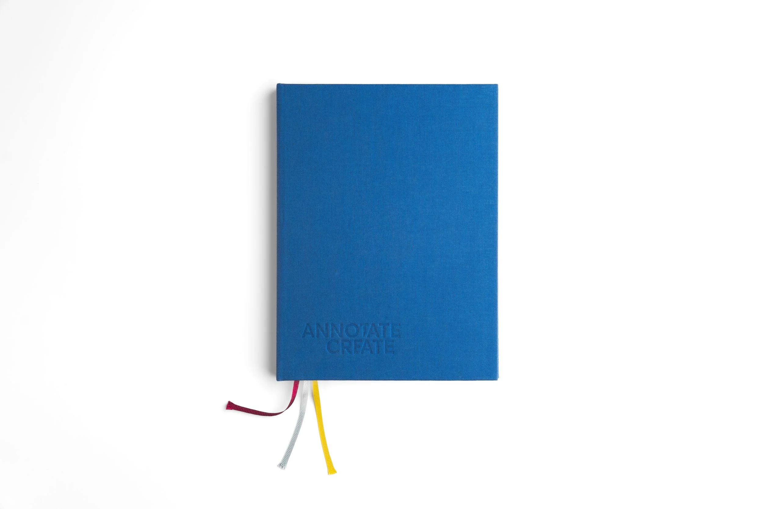 A blue notebook with the words 'Annotate, Create' embossed on the cover. It has yellow, white, and red ribbon bookmarks hanging from the bottom.